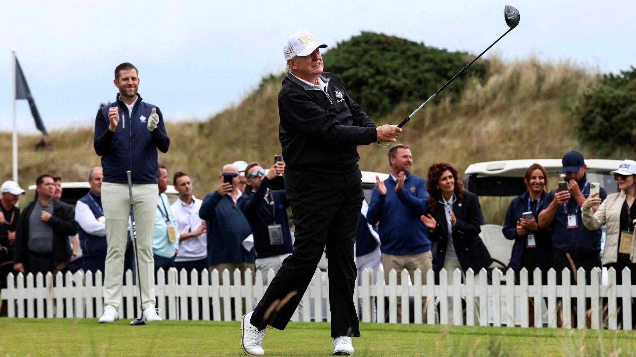 Donald Trump after taking a golf swing. His son Eric is applauding on the course, along with a number of spectators behind a small, white fence