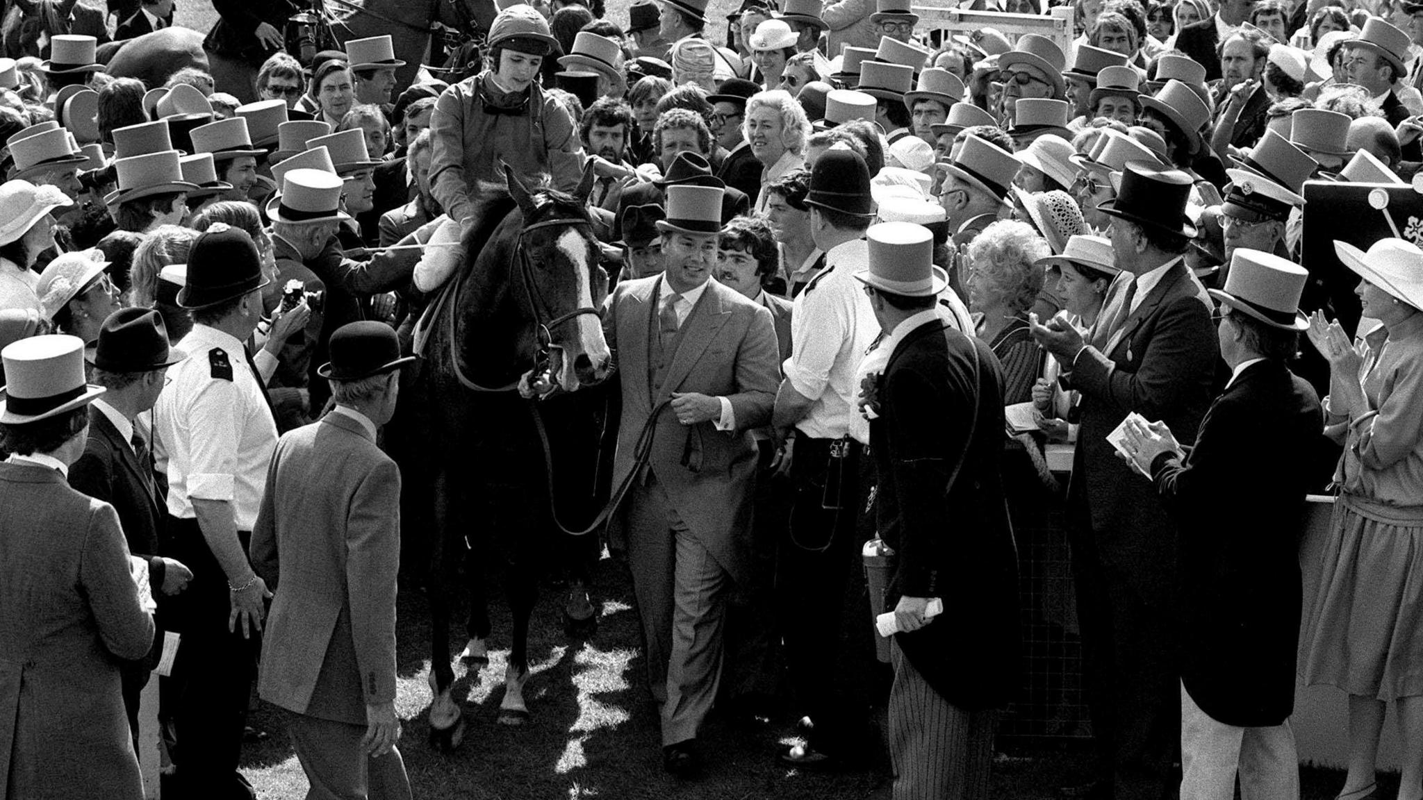 It is a black and white image of a crowd of dozens of people surrounding a horse with a white stripe on the front of its face. A jockey is sitting on the horse and a man wearing a top hat and morning suit is leading the horse through the crowd.