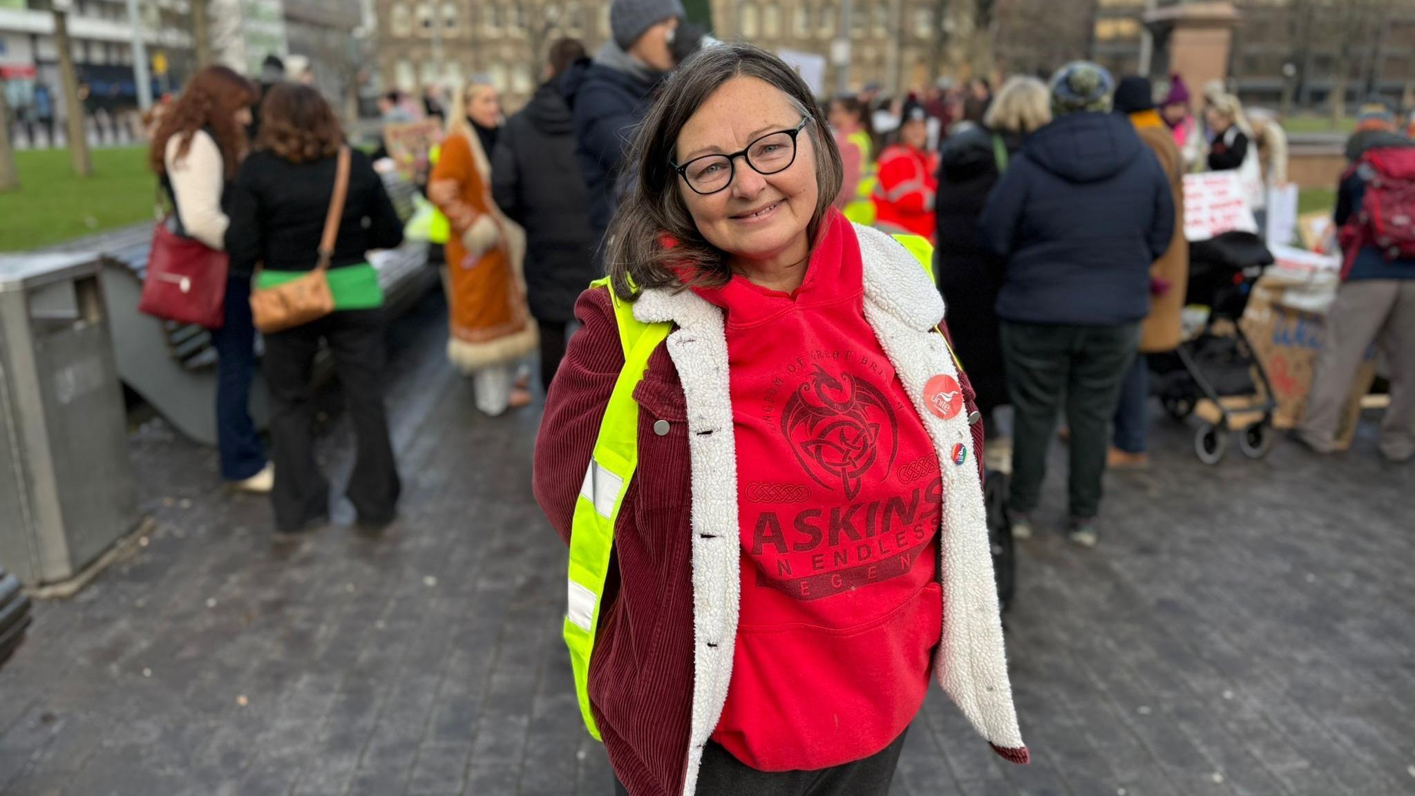 Liz Askins smiling at the camera with her hands behind her back. She has dark hair and is wearing a hi-vis yellow vest and dark glasses.. Protesters can be seen behind her.