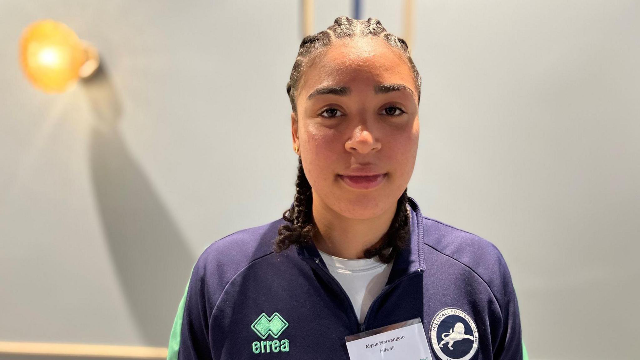 A face shot of Alysia Marcangelo, who is looking at the camera with a straight face. She is wearing a navy blue Millwall tracksuit top and a name badge.