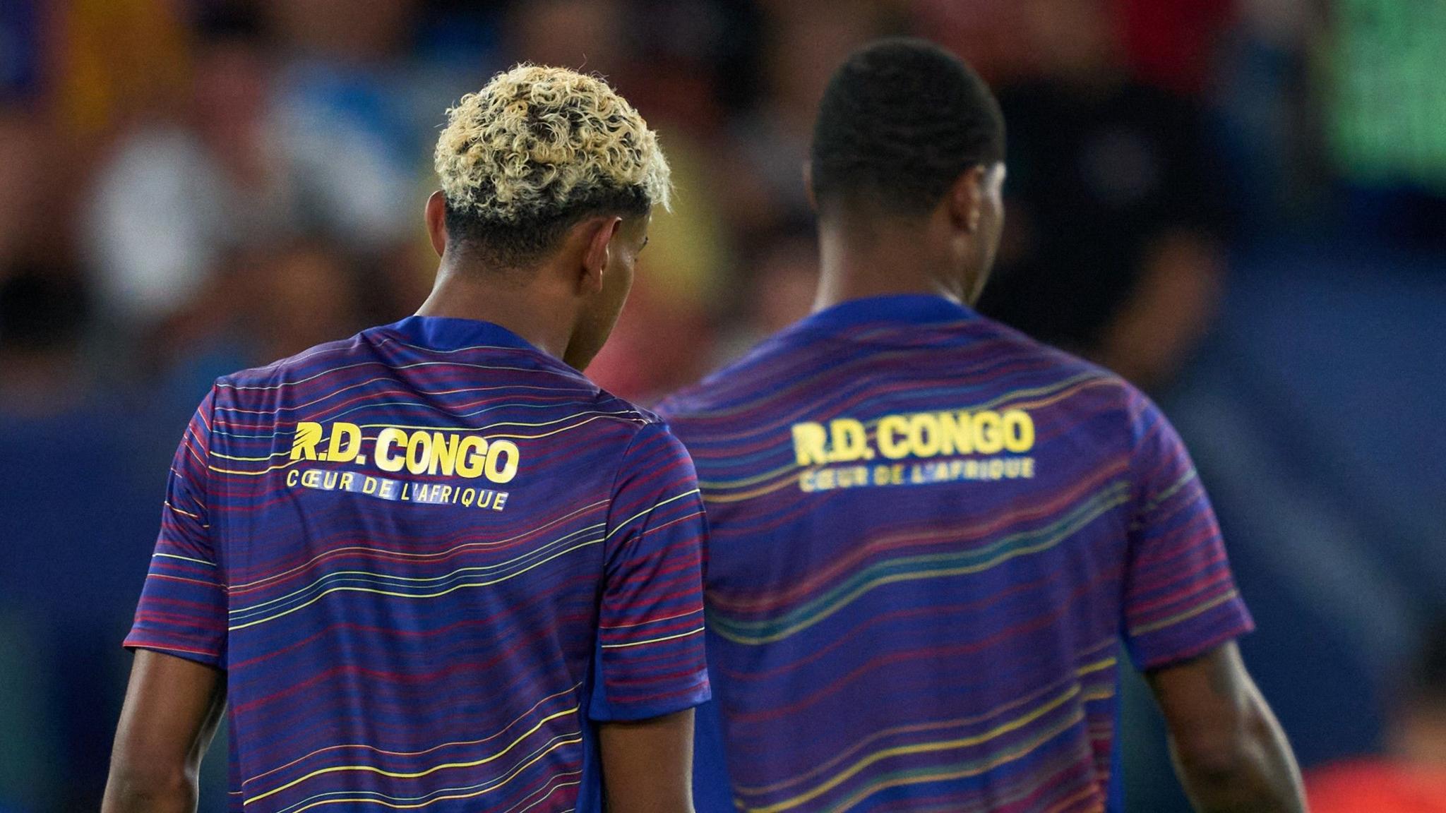 Barcelona players Lamine Yamal (left) and Marcus Rashford (right) are seen from behind during the warm up to a game. Yamal, with his bleached blonde hair, is nearer to the camera and fully in focus, while Rashford is slightly blurred. The pair are visible from the waist up, showing their purple training tops which feature wavy horizontal lines in various shades of red and yellow, and the phrase "DR Congo, the heart of Africa" written in yellow text in French across the upper back