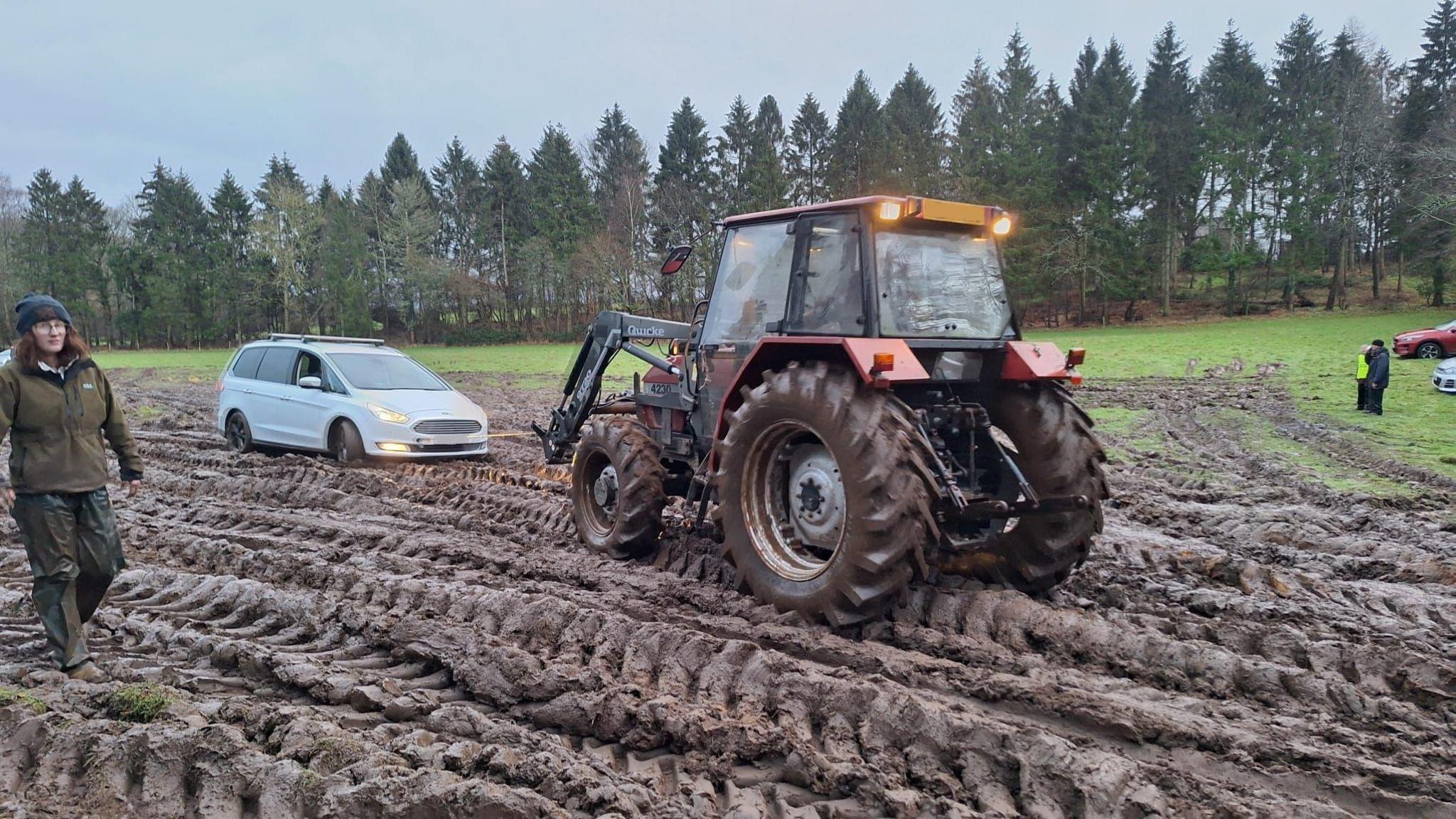 A tractor pulling a silver car over badly churned muddy ground, with a row of trees in the backgroud.