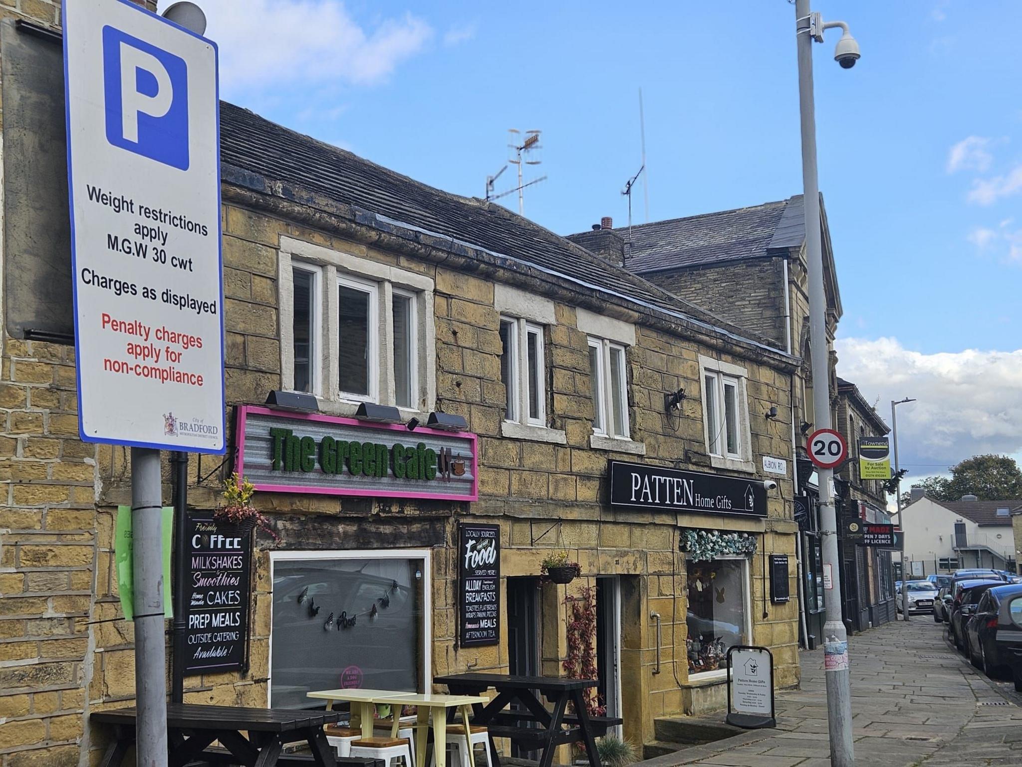 A street scene of a village high street with a view looking down a hill and the frontage of a cafe, a pub and other shops in view. In the foreground of the picture is a parking sign.