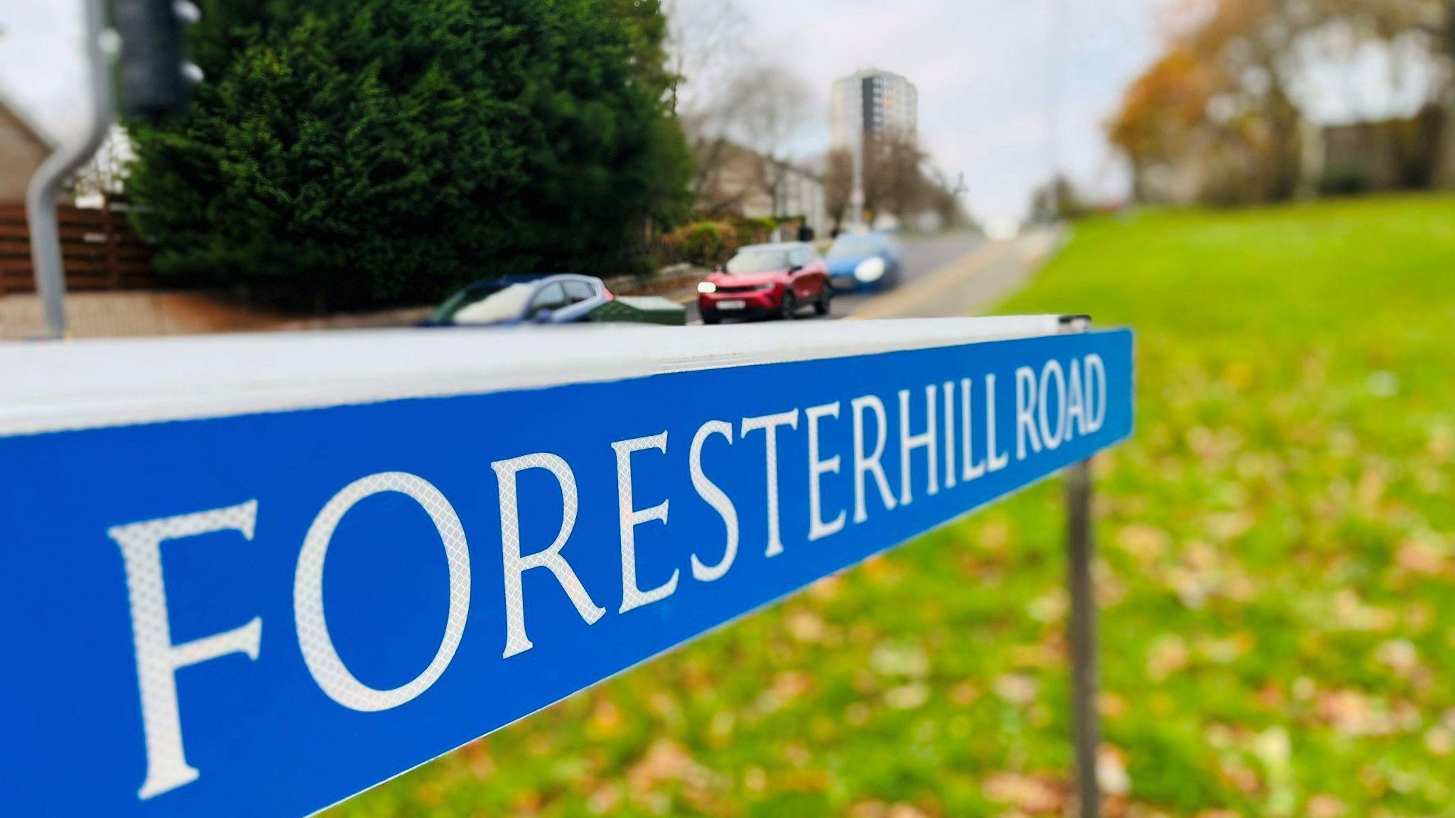 Blue road sign with white writing saying Foresterhill Road, on leafy grass, with cars and buildings in the background