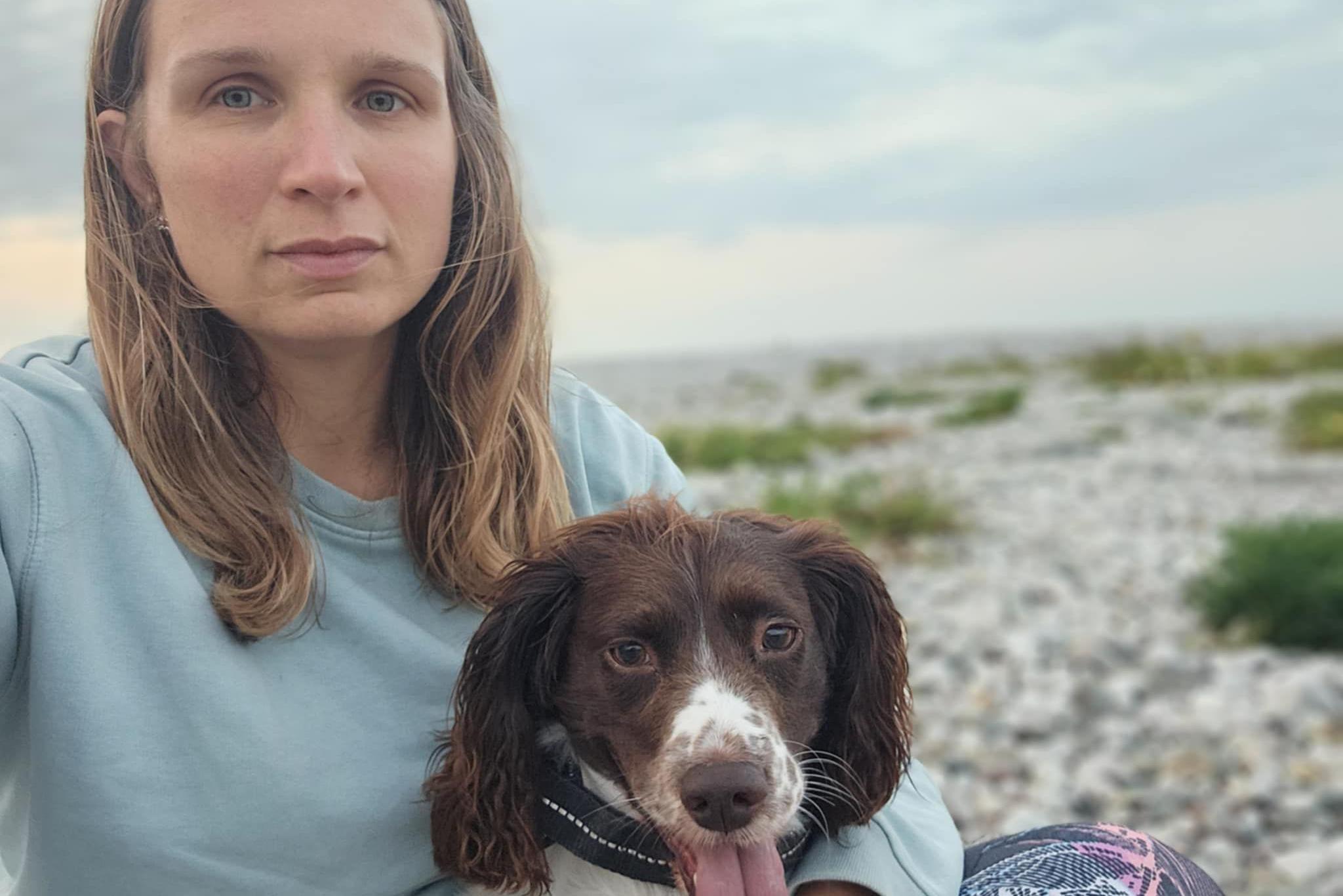 Laura, who was long blonde hair, wearing a blue sweatshirt, and holding a brown and white spaniel. They are sat on a pebbly beach with the sky and sea in the background. 
