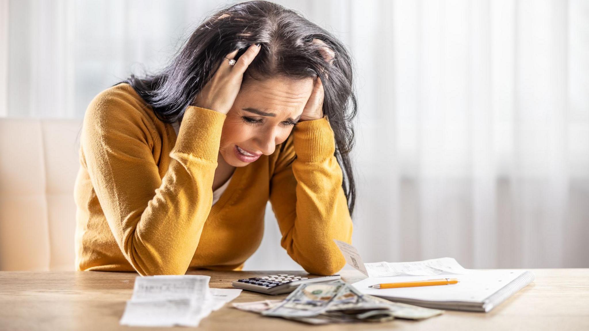 Brunette woman with yellow jumper has her head in her hands at the table over a pile of receipts and cash.