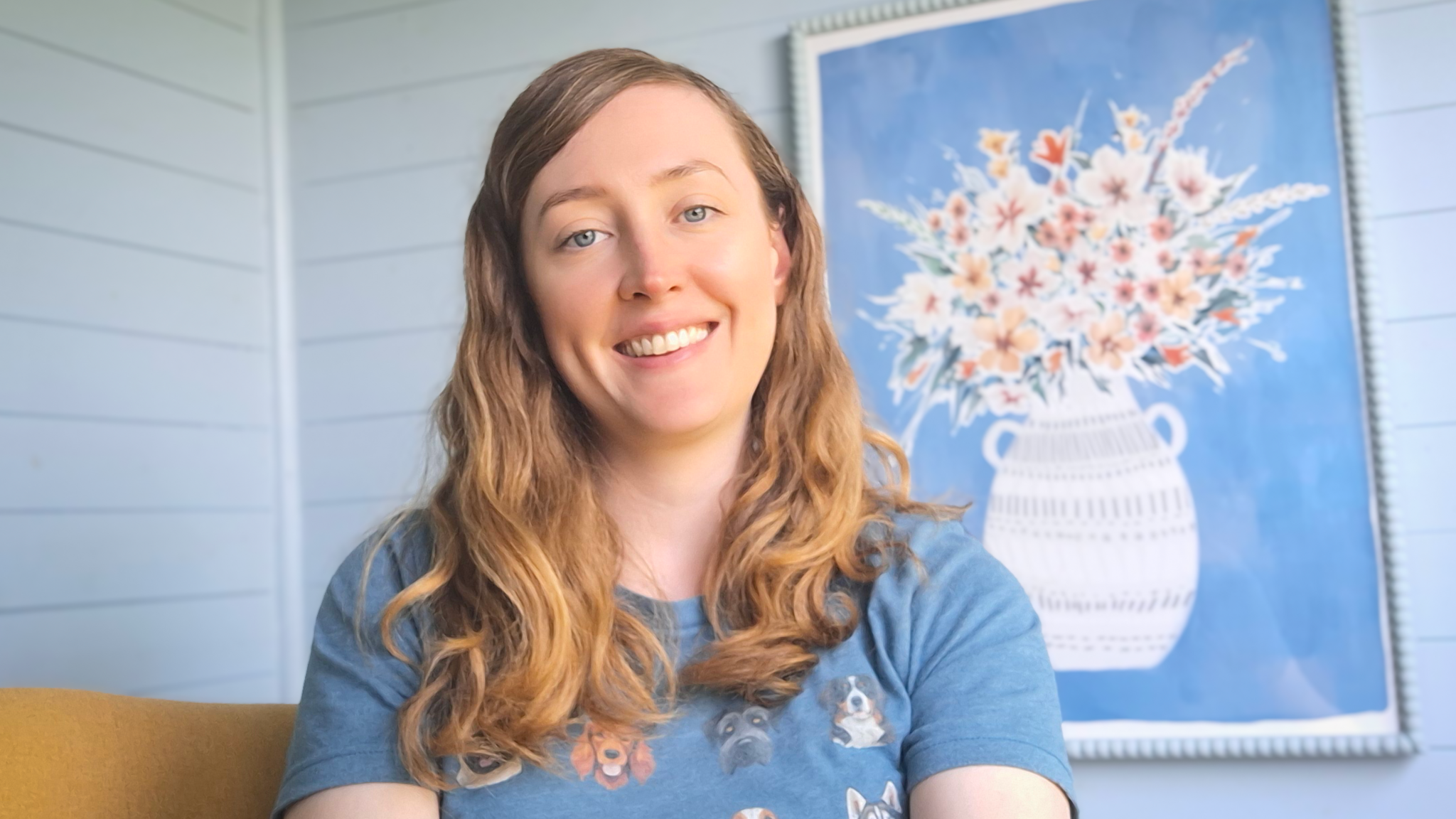 Sian Lawley-Rudd, a women with long blonde-brown hair smiles while sitting on a sofa in front of a painting of flowers. She is smiling and wearing a blue t-shirt with dogs on it.