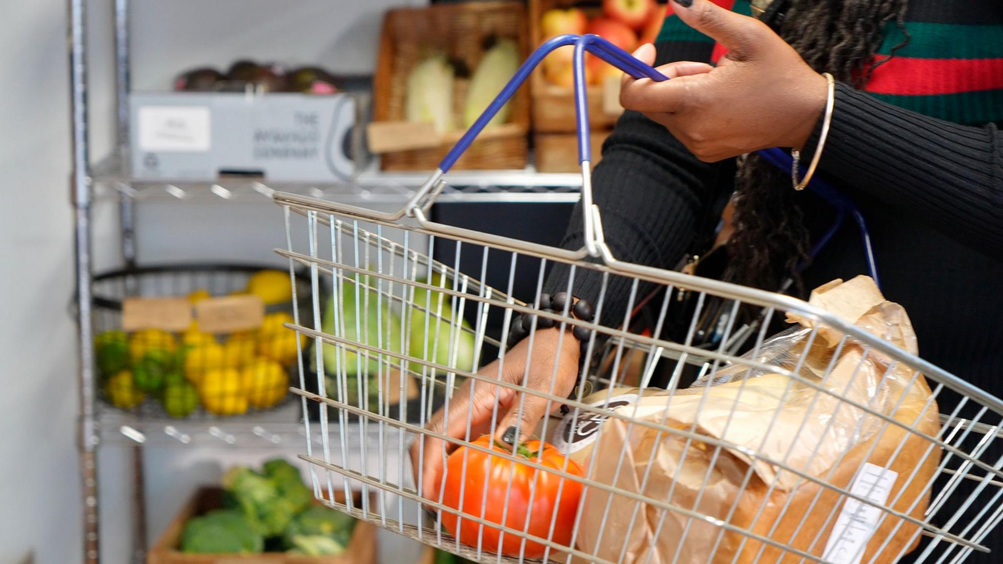 Close up of shopping basket with hand holding a large tomato and other items inside it