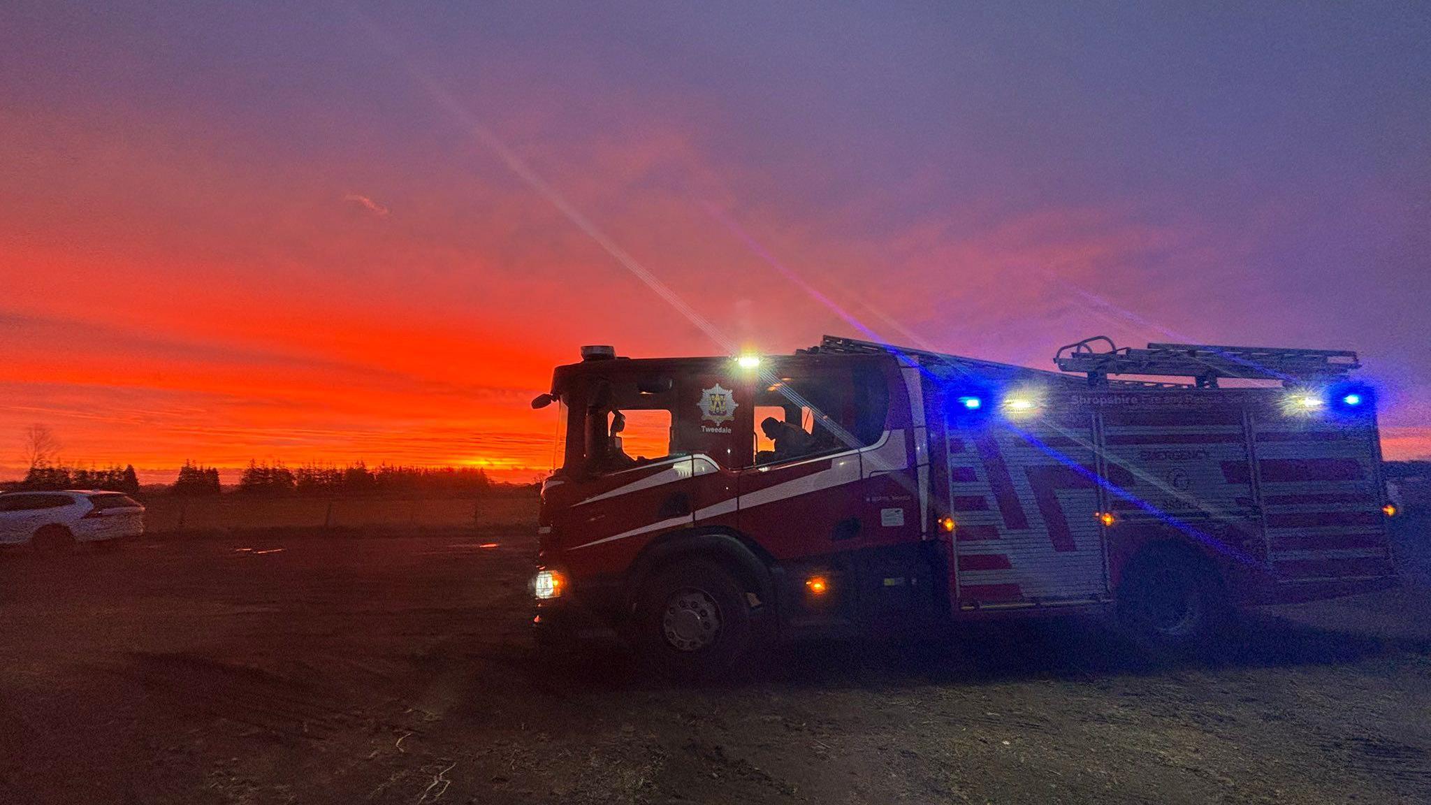 A Shropshire fire and rescue service fire engine with a person inside. its blue lights are flashing. it is parked in an open space with a sunrise behind it that is orange, pink and purple.