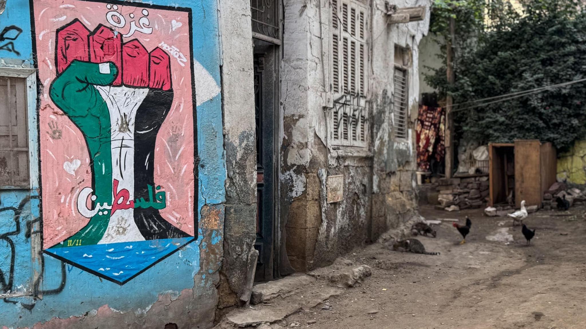 Street art in Cairo shows a raised fist painted in the green, white, red and black colours of the Palestinian flag in solidarity with Gaza. Buildings on the small, dirt-track street appear to be in bad repair. In the background are few chickens.