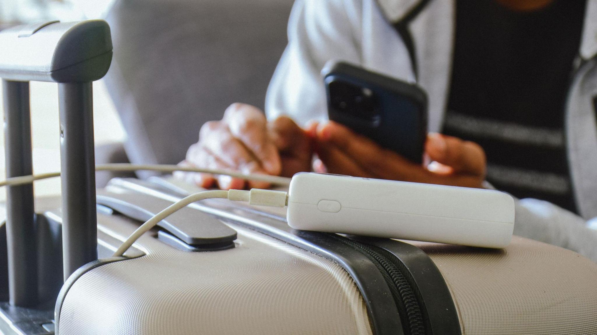 A white portable charger rests on top of a cream carry-on suitcase. It is plugged into a white cable, which is attached to a black iphone being held by a man.