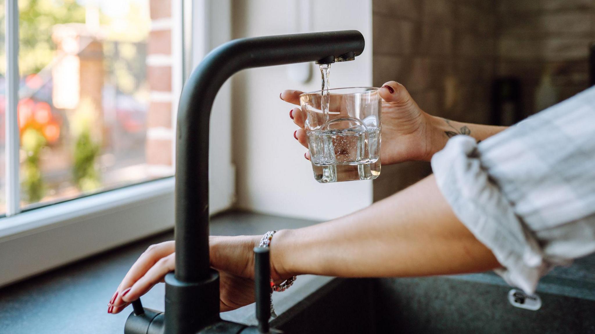 Someone pouring water into a glass from a black tap. You can only see their arms and hands. There is a window in the background.