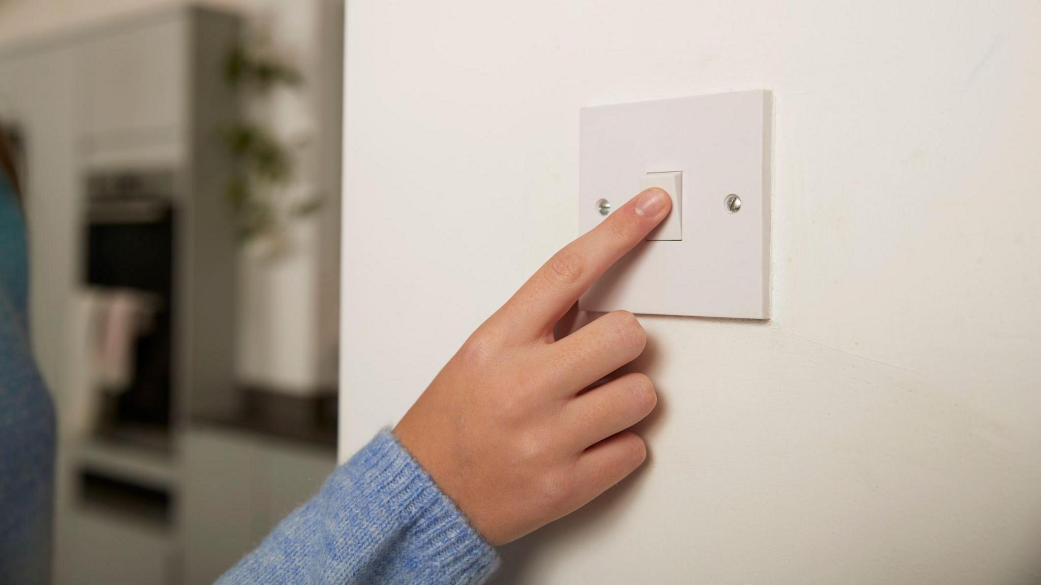 A hand switching off a light switch. The person is wearing a blue knit jumper and the white plug is on a white wall. You can see the kitchen in the background. 