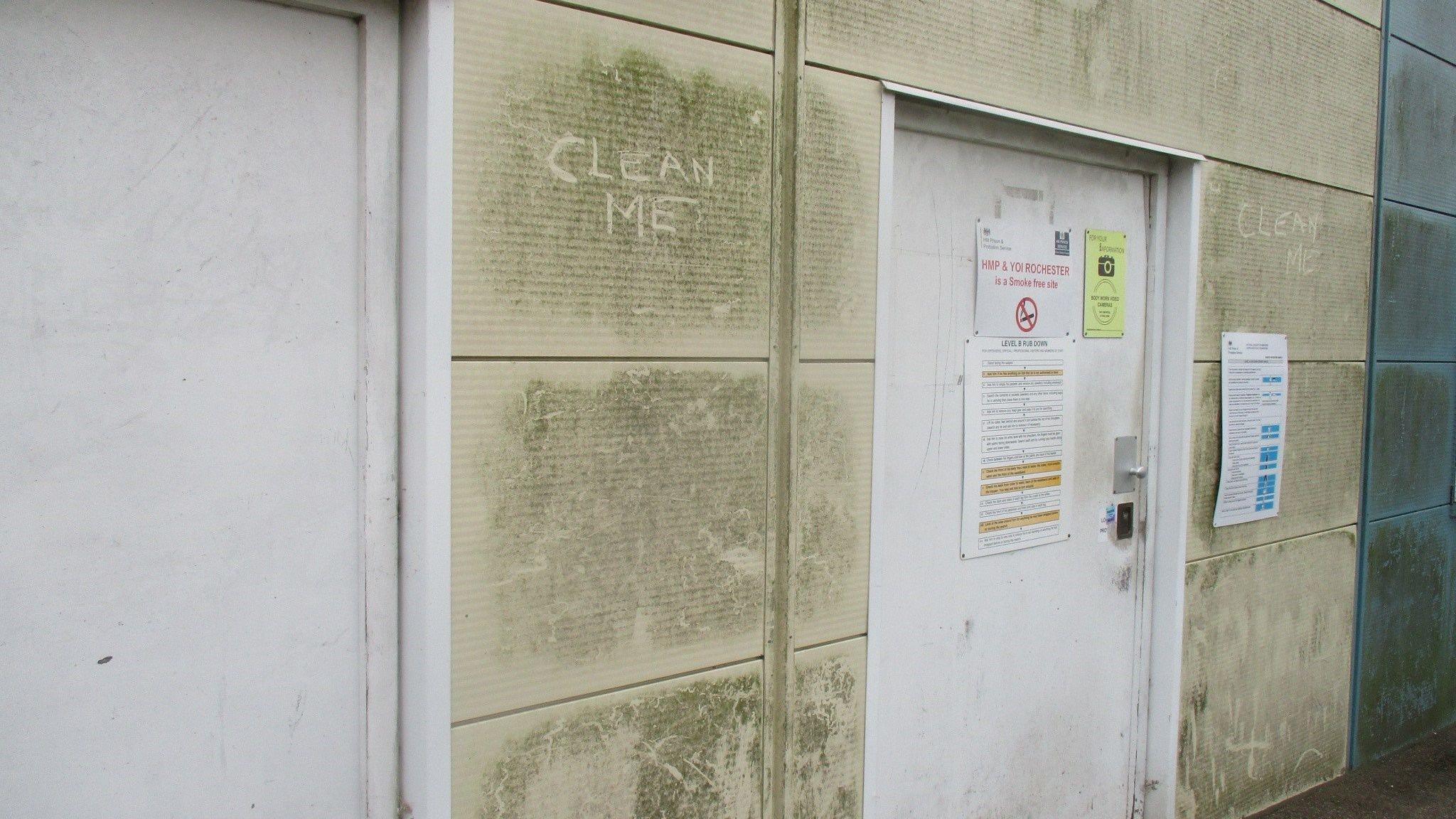 A dirty outside wall is seen surrounding a white door with signs on it. In the dirt on the wall, someone has written "clean me" in capital letters in two places.