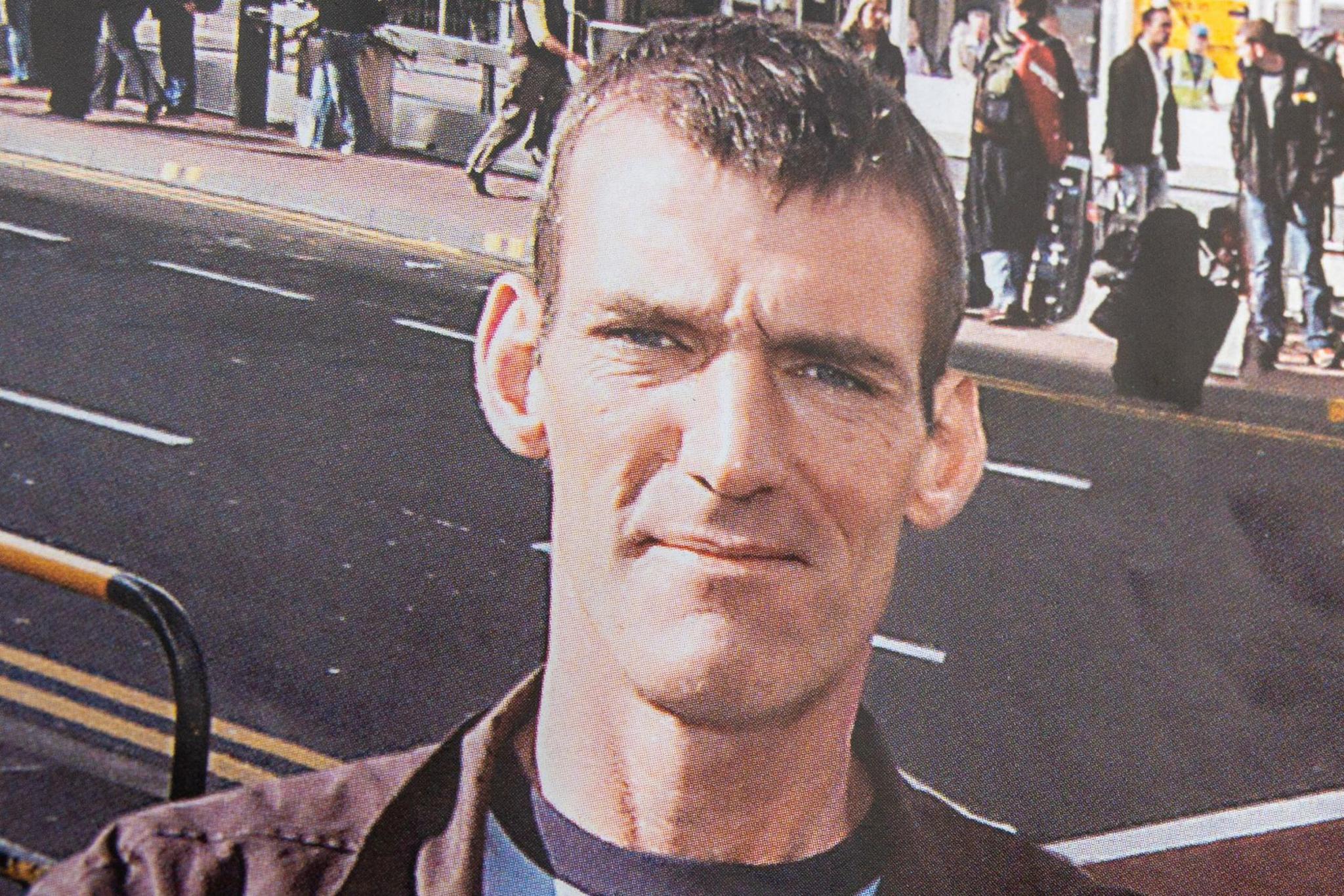 A man with dark cropped hair stares at the camera. He is wearing a brown jacket and a blue shirt. There are airport passengers in the background.