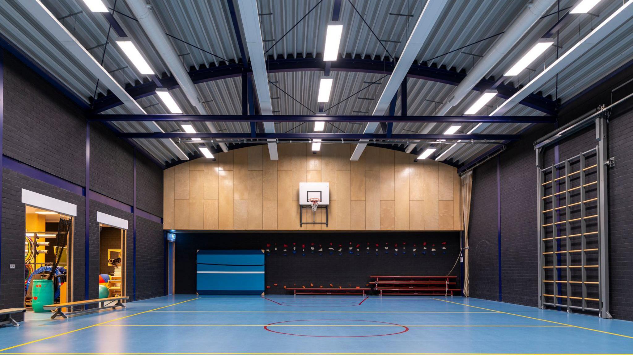 The inside of a school gym, with climbing bars on the right hand side, packed away, and a basketball court and hoop taking up most of the hall.