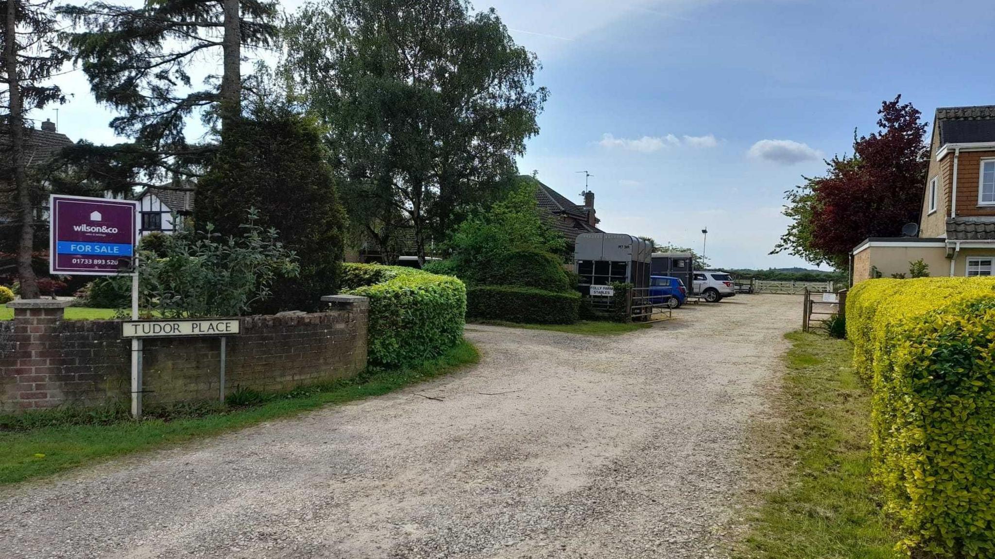 A gravel path with hedges and houses on either side. There is a 'For Sale' sign outside the nearest home above a road sign which reads 'Tudor Place'. There are also several trees and a horse box visible.