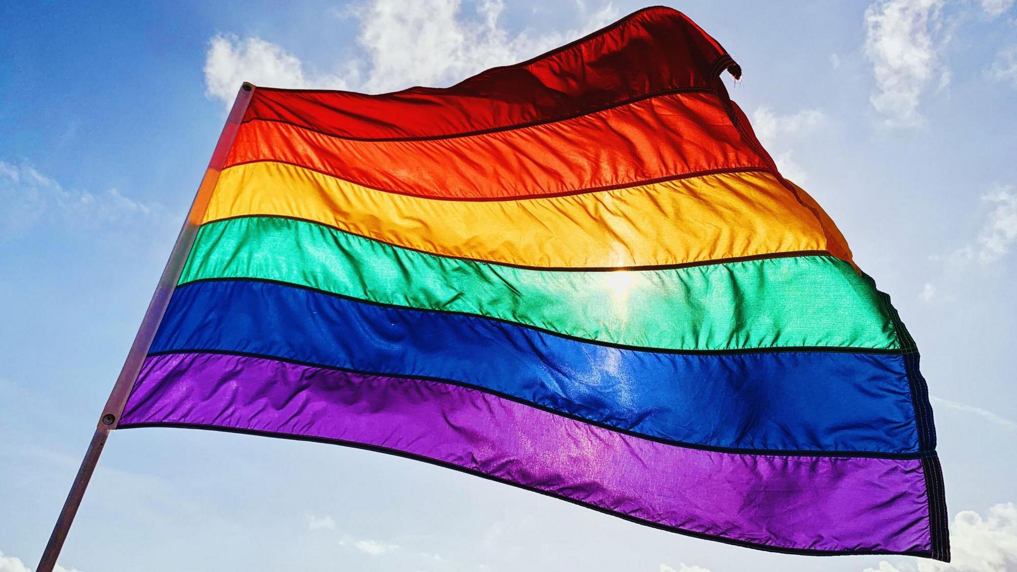 A rainbow flag flaps in the wind with the sun behind it. 