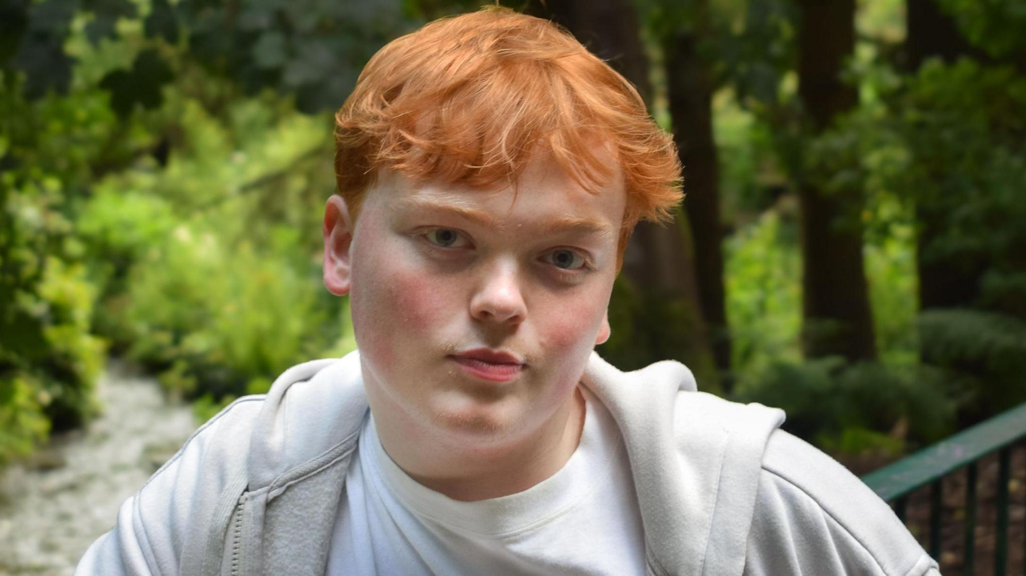 Mr Horne is wearing a white top and light jackets while standing in font of trees with lots of green leaves. He is looking directly at the camera and has ginger short hair.
