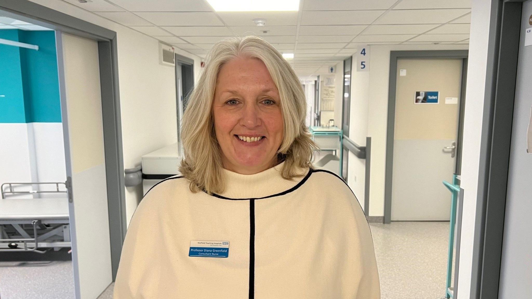 Professor Diana Greenfield stands in a hospital corridor wearing a white jumper with a black stripe down the front.  She has grey hair and is wearing a hospital name badge.