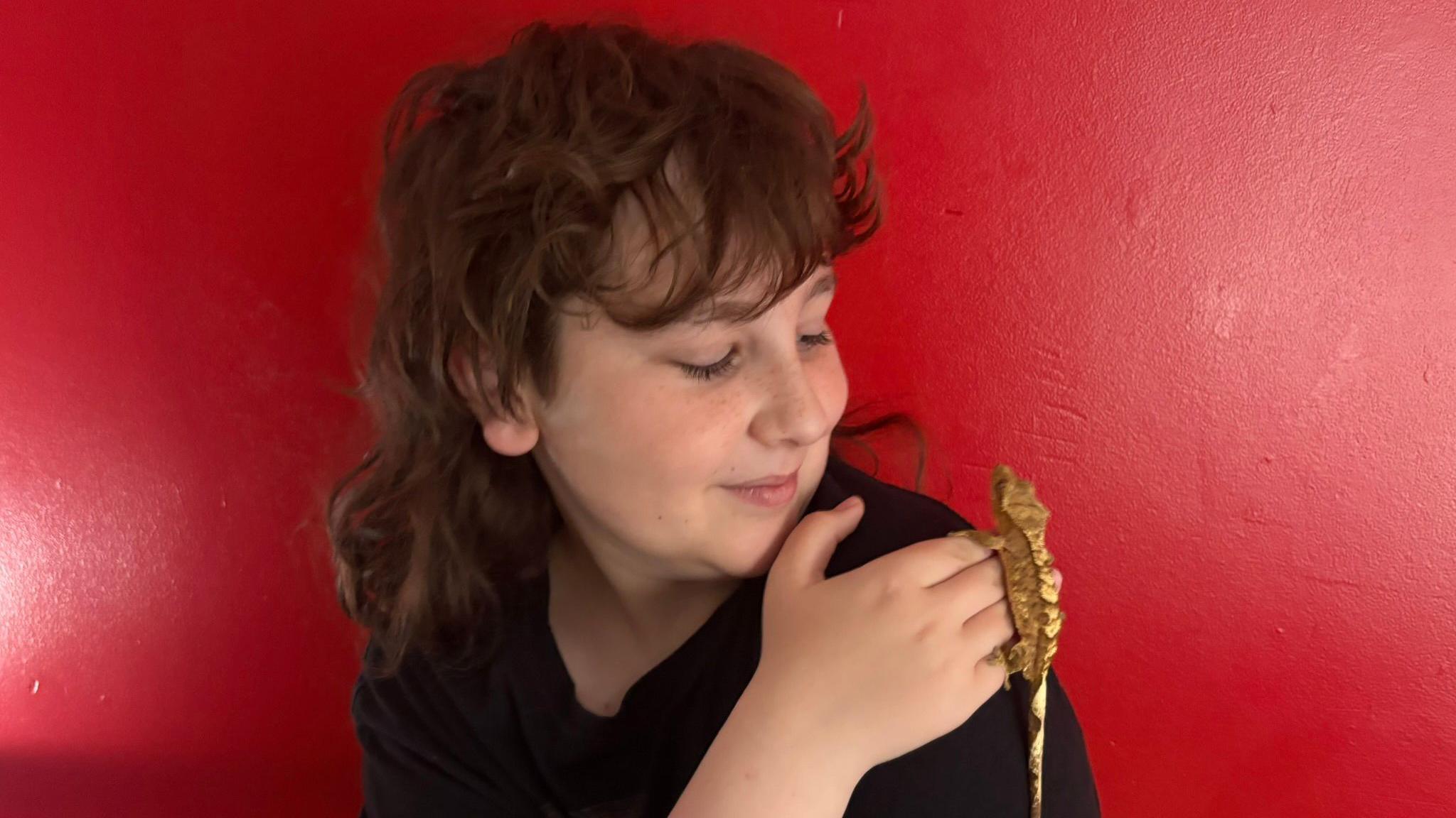 A boy with long brown hair and a fringe is wearing a black top and has his hand on his shoulder which he is looking at. A sandy coloured gecko is resting on his hand. The background is a red wall.