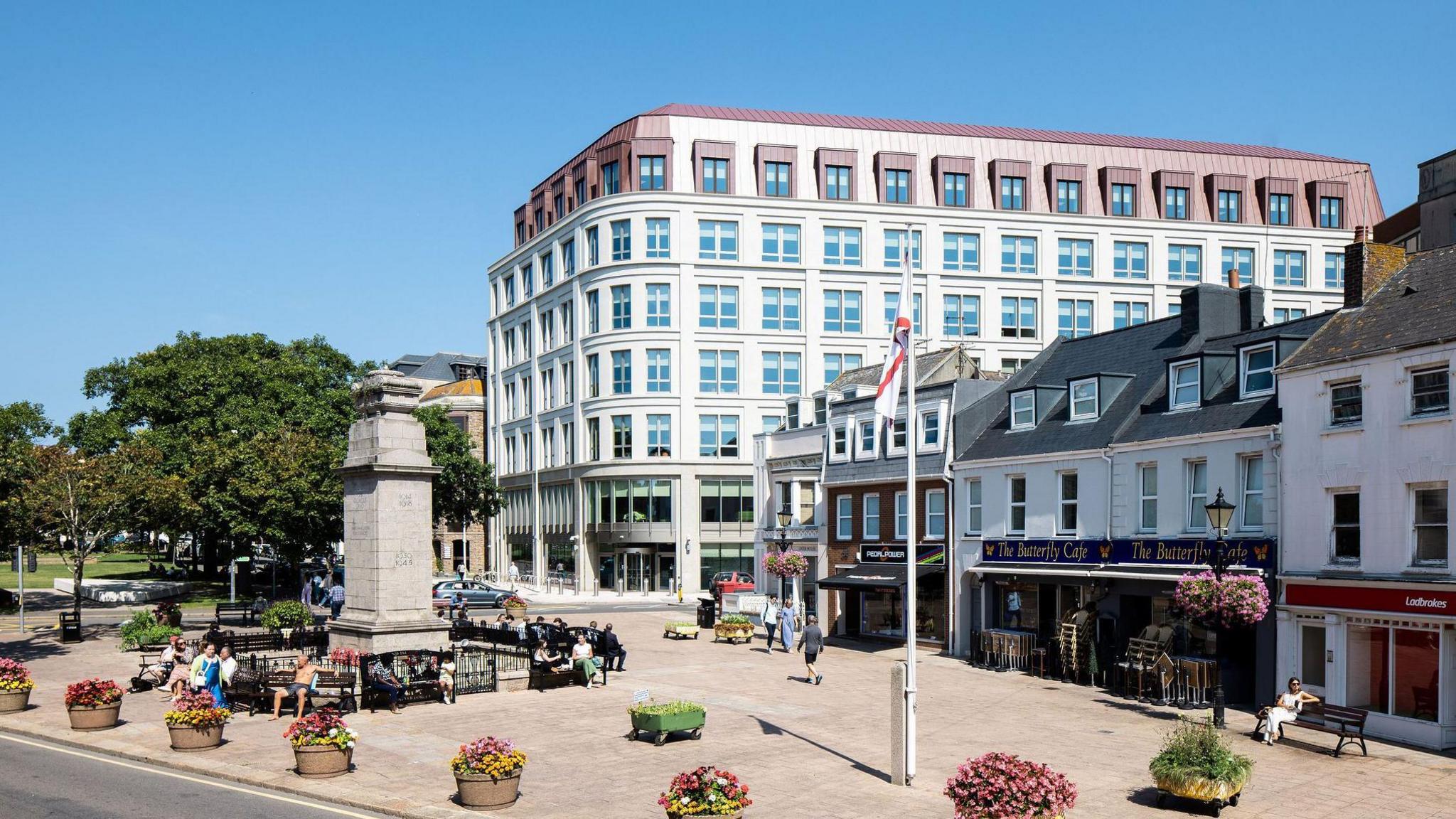 A large white building with many windows sits at the corner of Union Street. The building is new. It sits behind a row of older properties and a monument. There are green trees to the left of the building. 