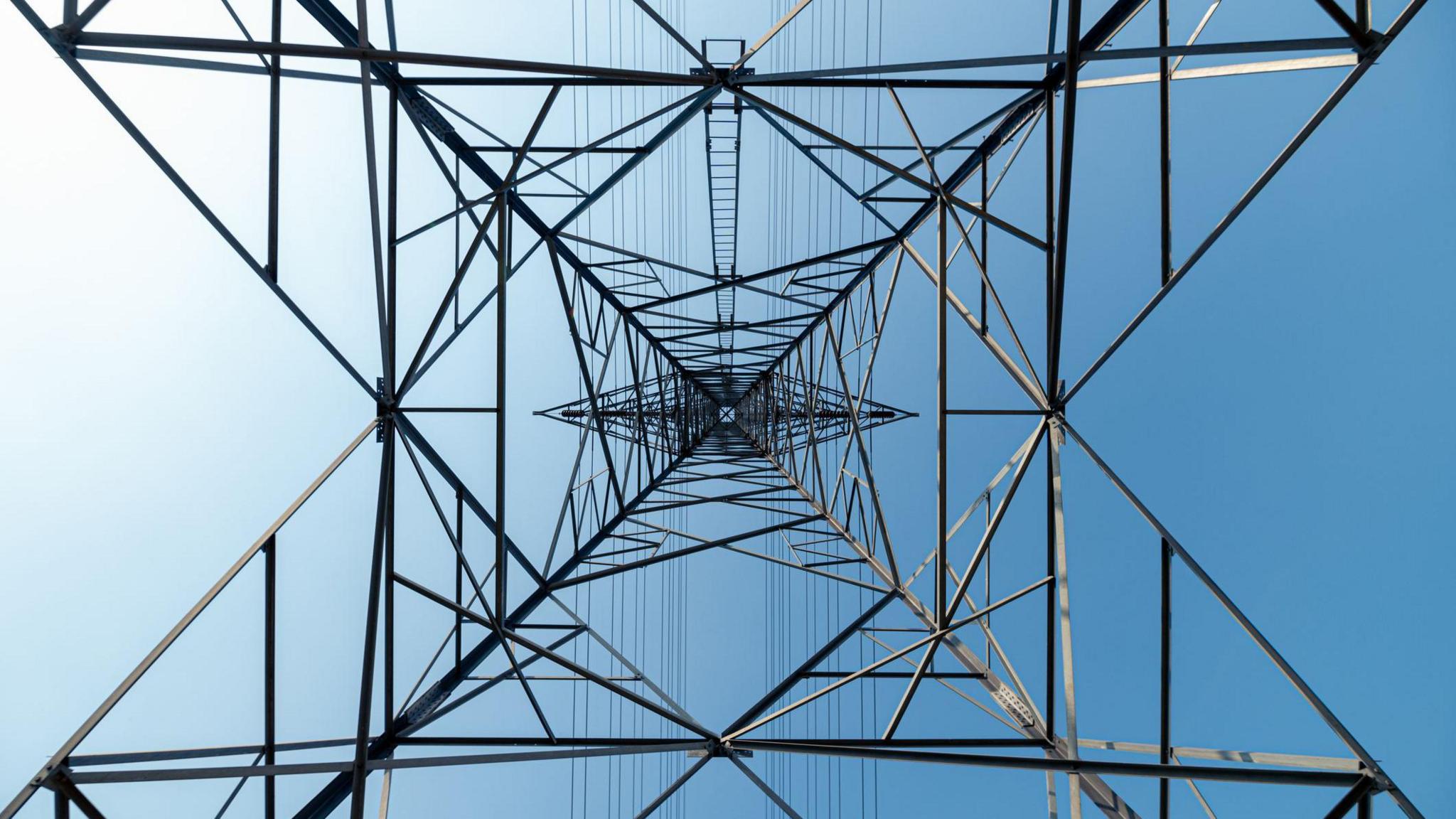Library image of an electricity pylon. The camera is pointed upwards from the base of the structure. It is made of metal and has wires strung across horizontally. Blue sky can be seen in the background.