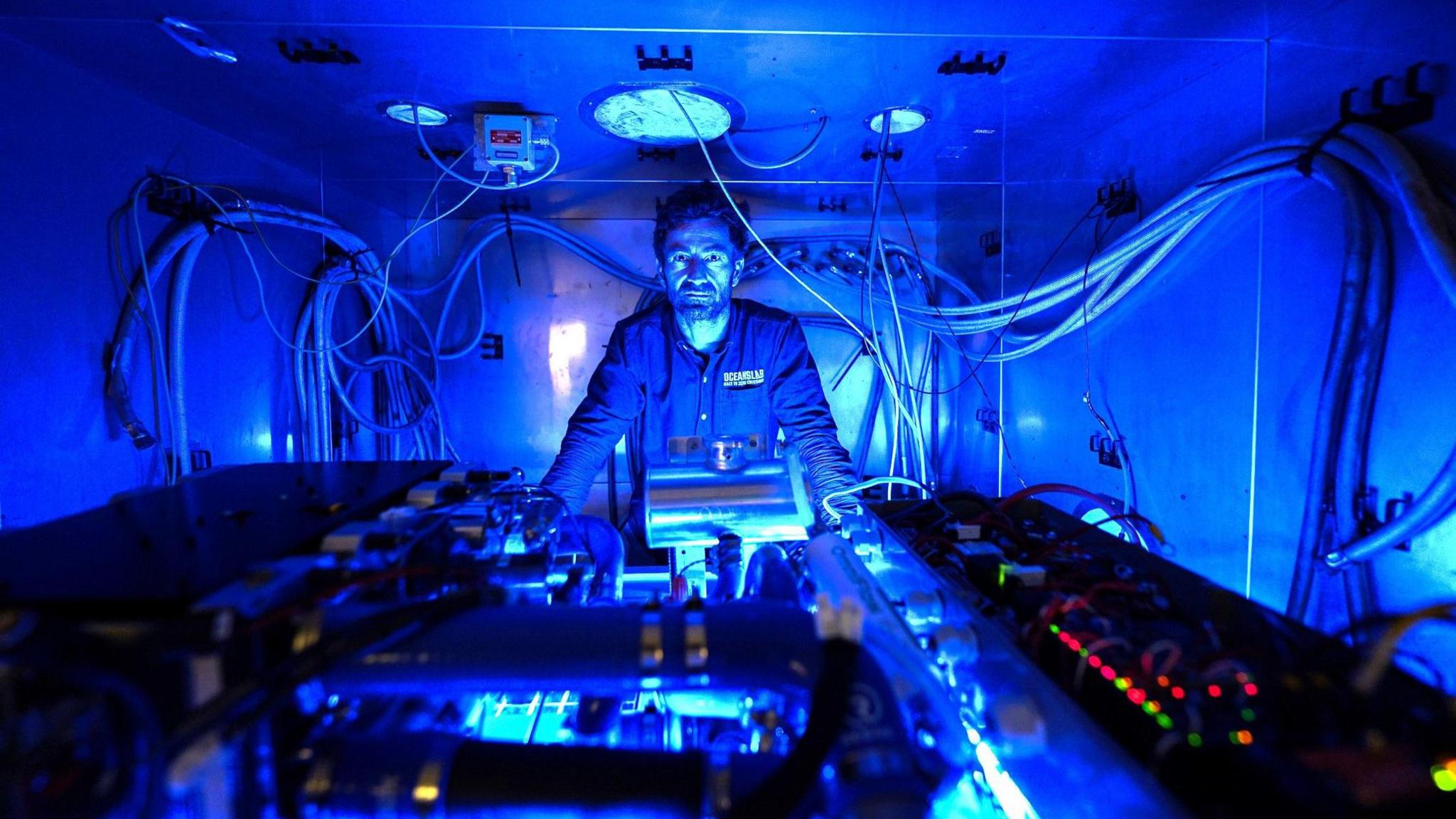 A heavily blue image of a man in overalls in a small metallic room with a hydrogen engine in front of him.