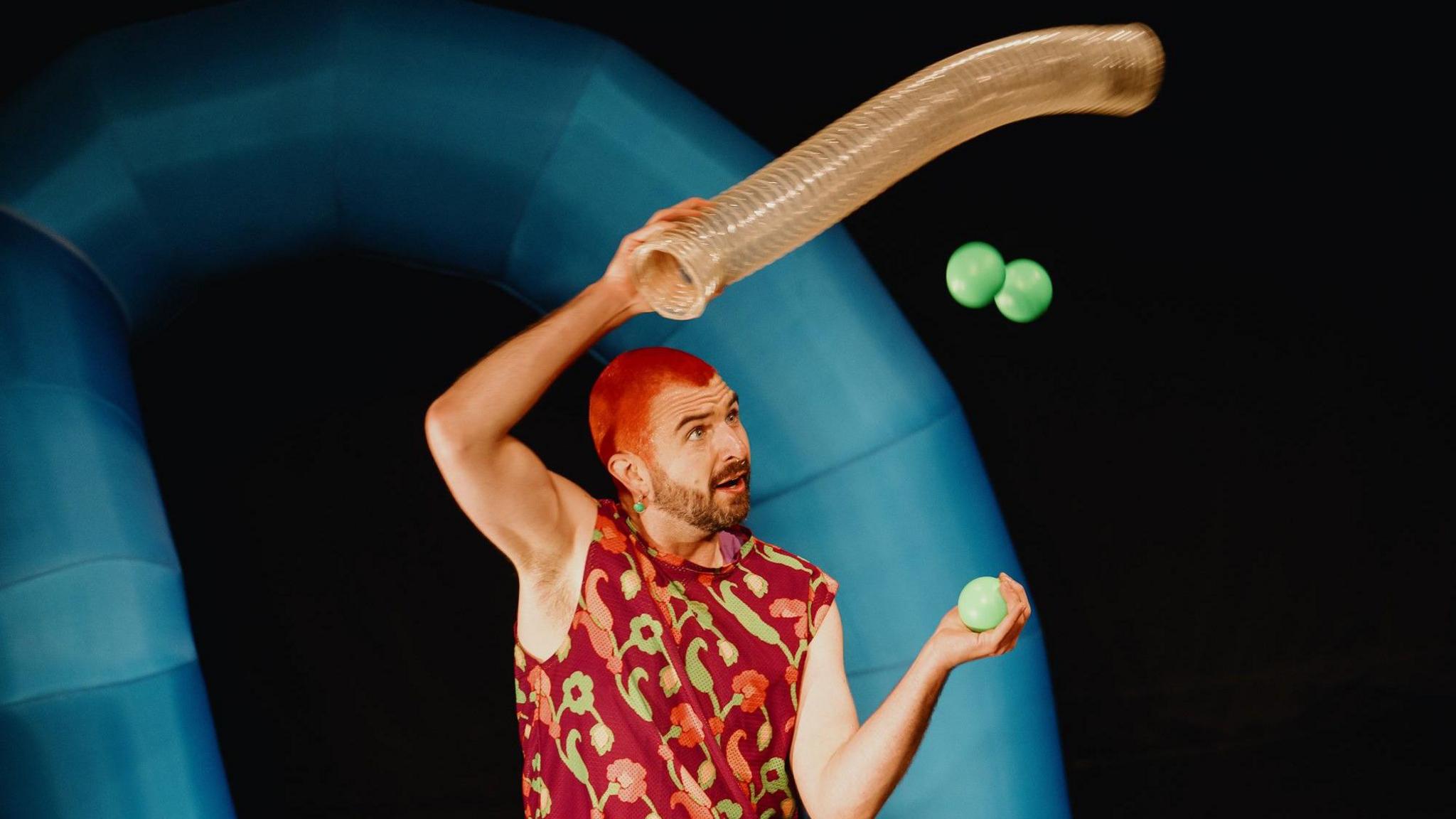 A man with red hair juggling on stage. He is wearing a red vest top with a red and green floral pattern.