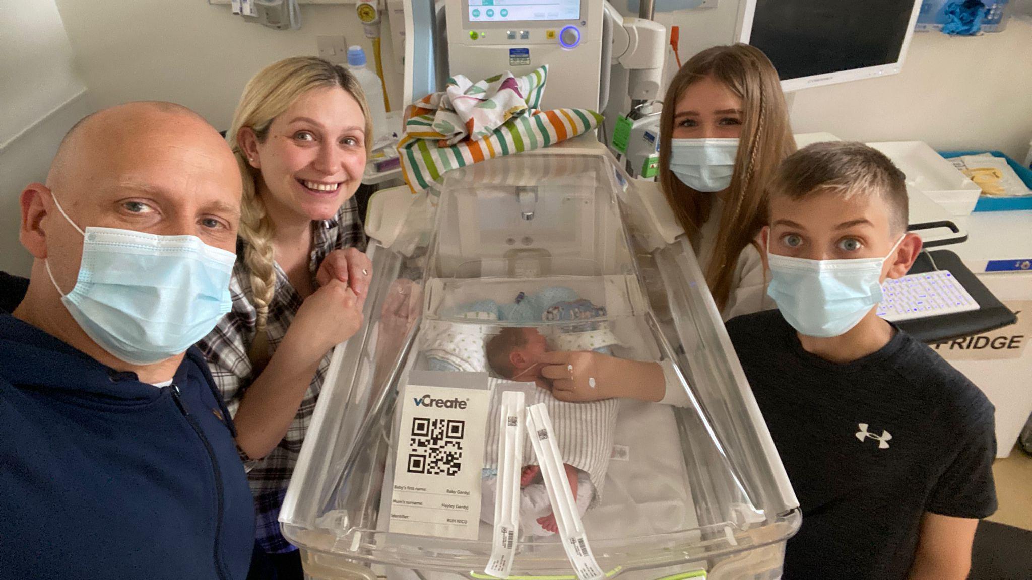 A baby is lying in an incubator in hospital. His two siblings, pictured on the right, are wearing Covid masks. His mum is wearing pyjamas and smiling to the camera, his dad, on the left, is also wearing a Covid mask.