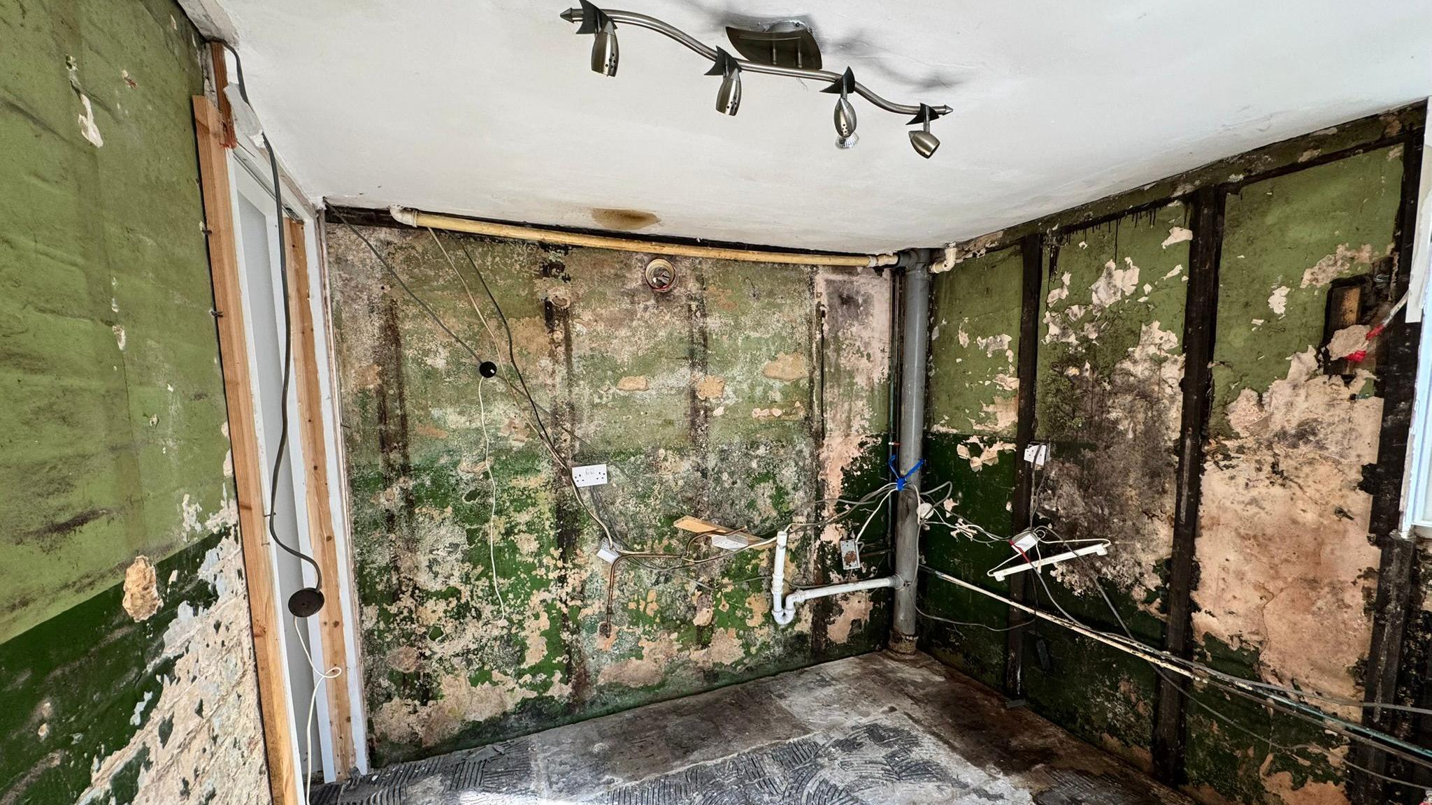 A room seen being gutted. The walls are green painted brick with many scraped and marked. There are sockets and wires dotted around. The floor is a layer of underlay and various pipes can be seen running vertically and horizontally. There is a smart silver set of spotlights on the ceiling.