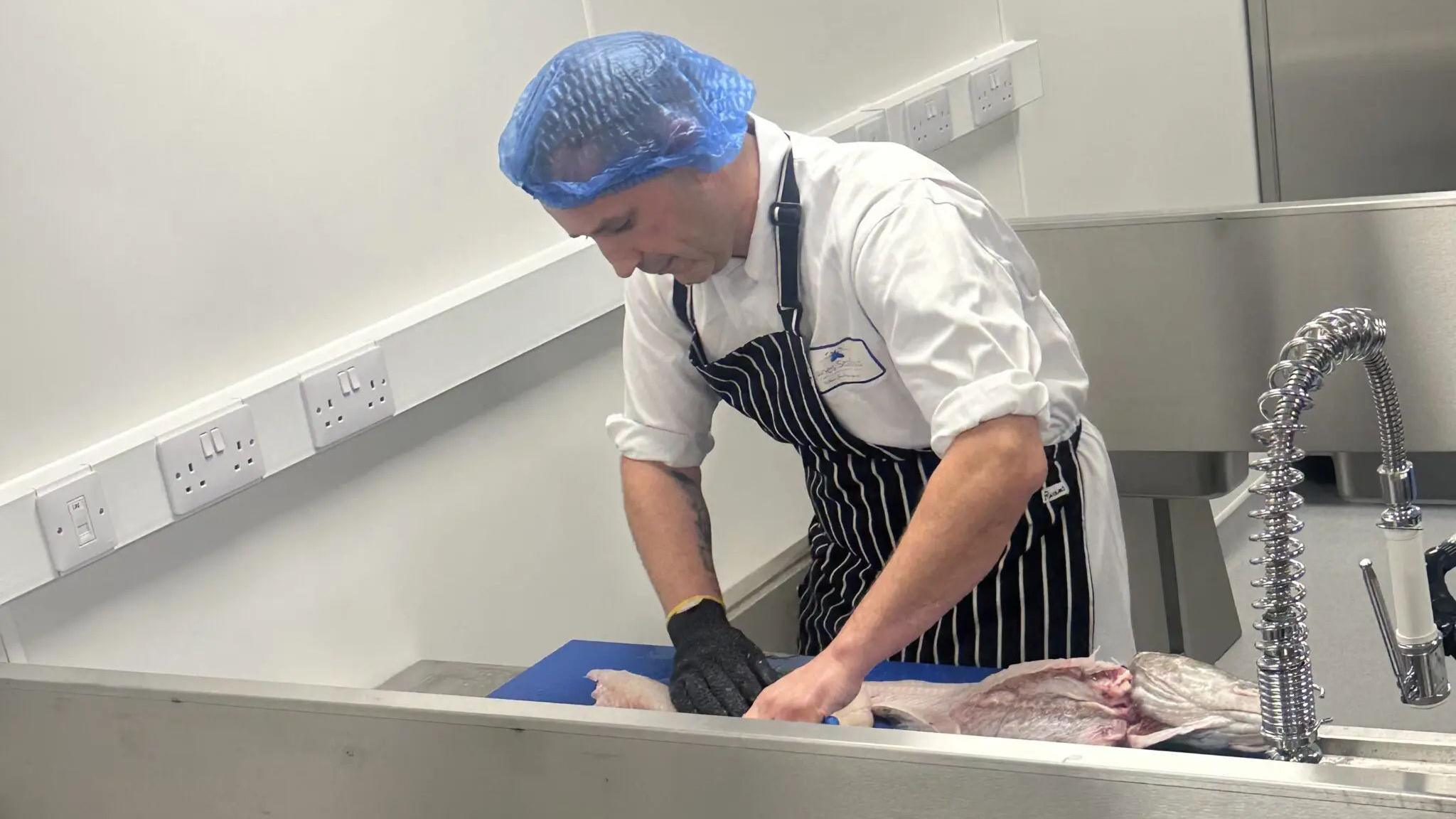 A man in an apron and a hair net, wearing a white chef's outfit, prepares a fish for cooking in a stainless steel kitchen area. 