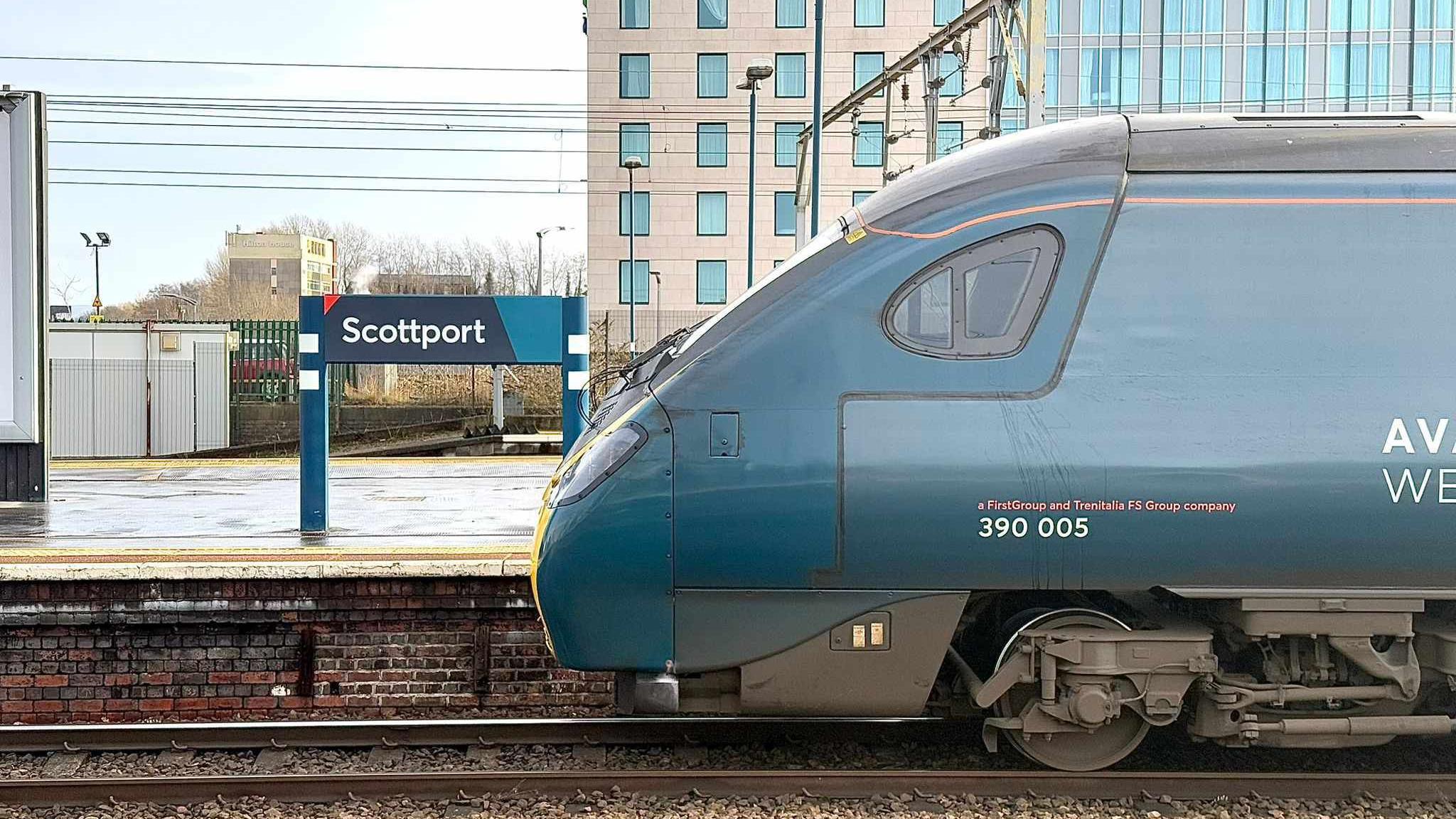 A blue sign for Stockport station, re-written to say Scottport in white lettering, erected outdoors on a train station platform, with a train car seen on railway tracks in the foreground. 