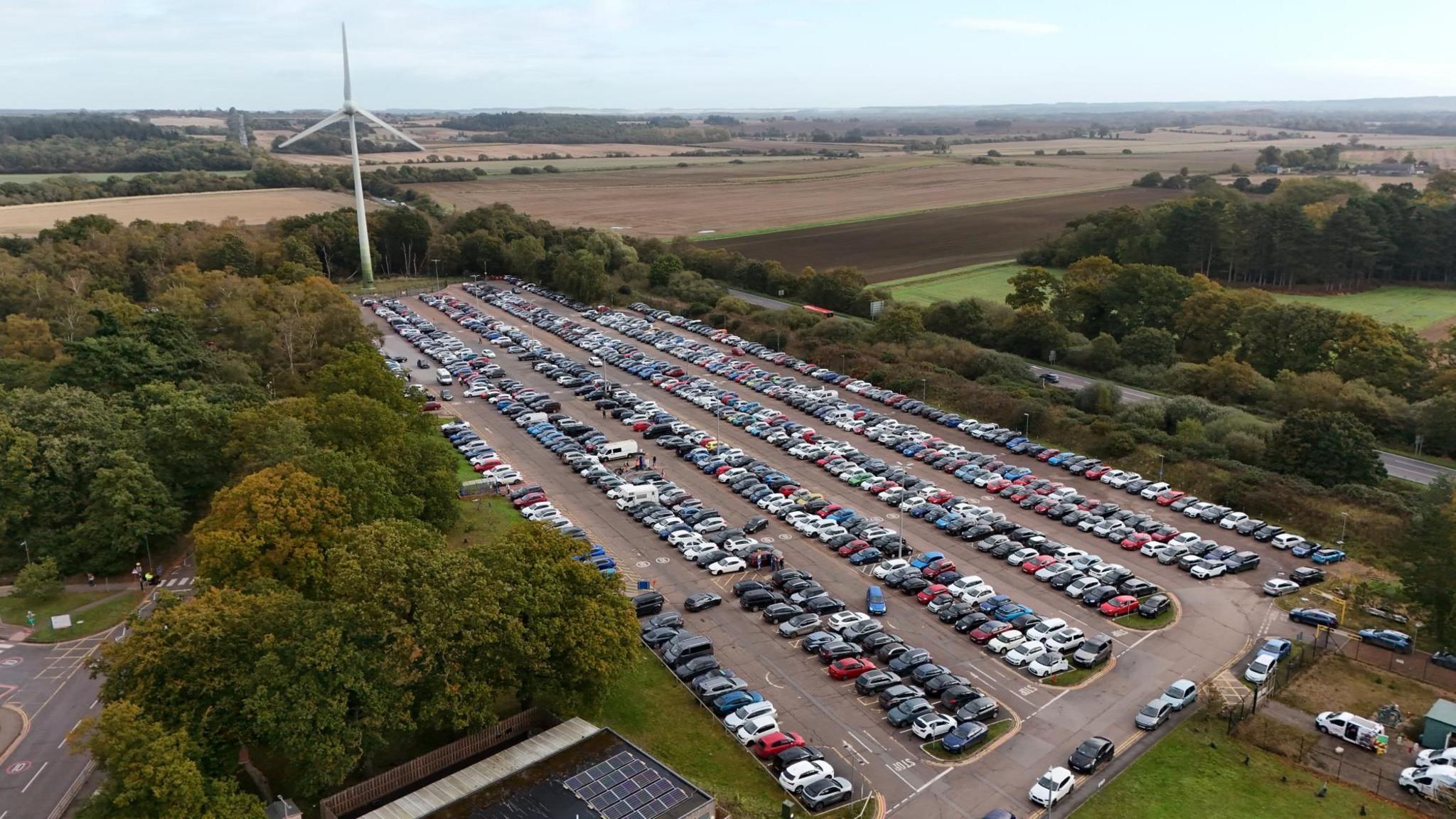 An aerial view of the car park of the Queen Elizabeth Hospital in King's Lynn which is full of cars. There is a wind turbine at one end of the car park and farmland surrounds the hospital