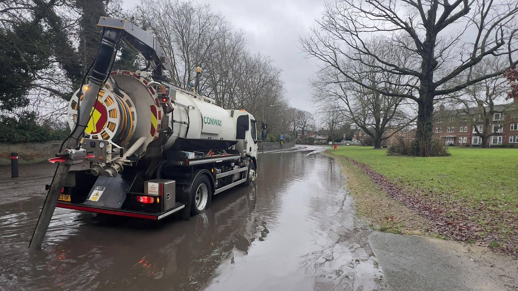 A white road tanker with a tube attached to the back of it. The lorry is parked on a flooded road with a patch of grass and trees to the side.