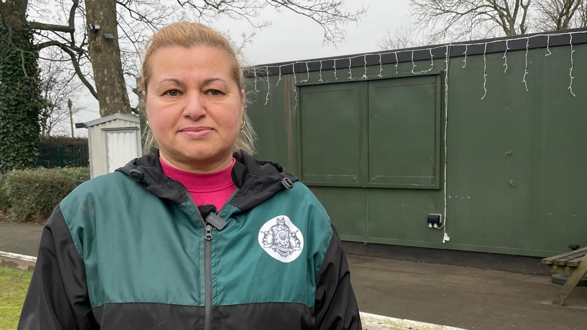 A woman with tied back blonde hair wearing a pink top and a green and black waterproof jacket stands in front of a green shipping container on a bowling green.