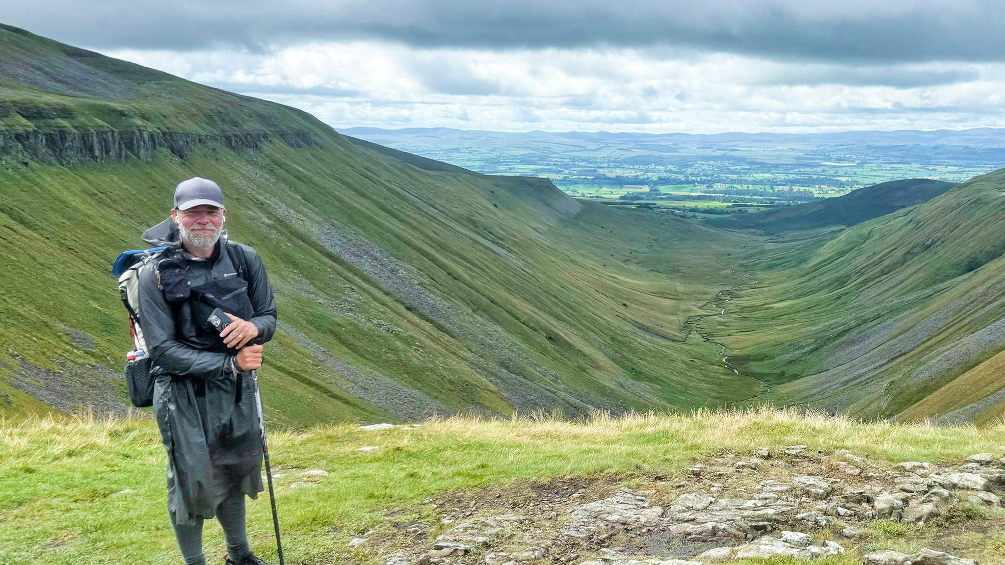 Mr Neal poses at the top of a ravine. He wears a cap and dark coloured walking clothing along with a walking stick.