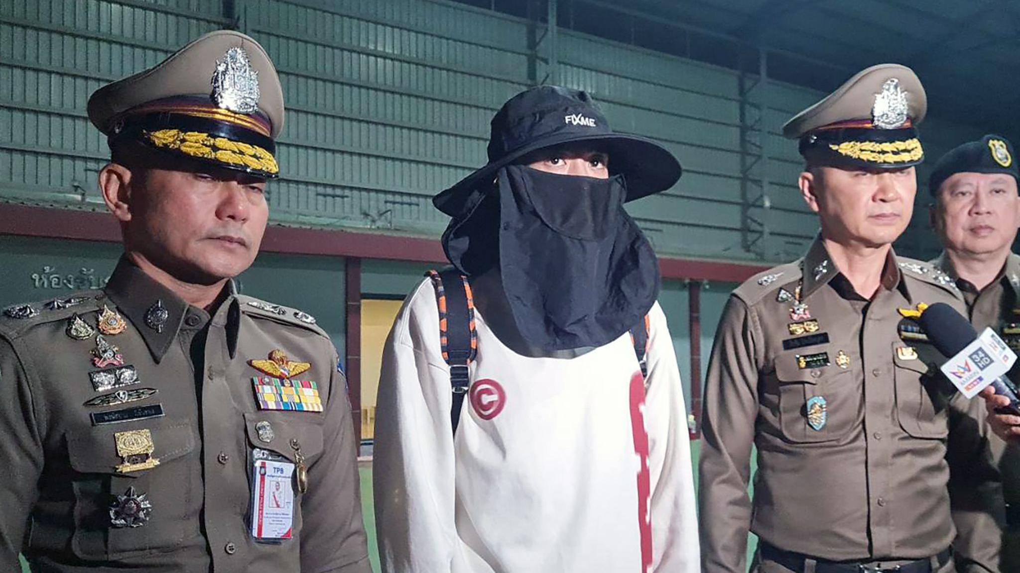 Wang in a white sweater, wearing a black hat and a black face mask, flanked by two Thai police officials