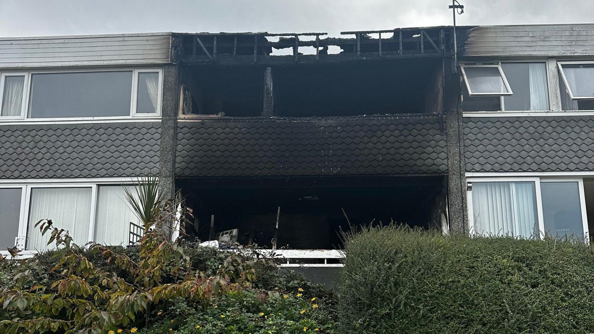 A late 20th Century terraced house of two storeys with a blackened burned out frontage with black soot staining the tiling on the front of the property and the roof pockmarked with holes and scars of the fire visible on the neighbouring properties either side.