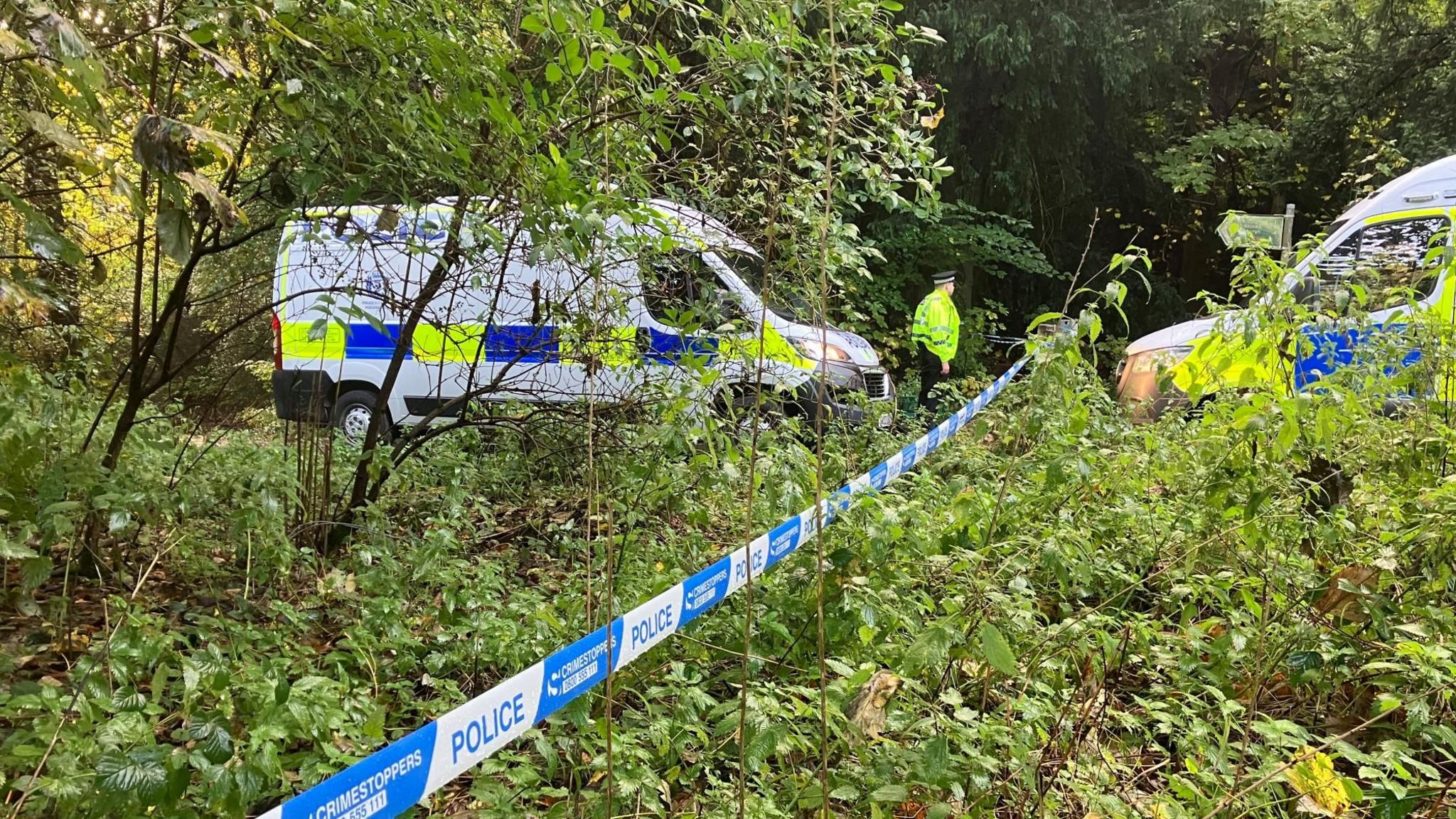 Two police vans facing each other in a wooded area, separated by police tape. There is a police office in front of one of the vans. 