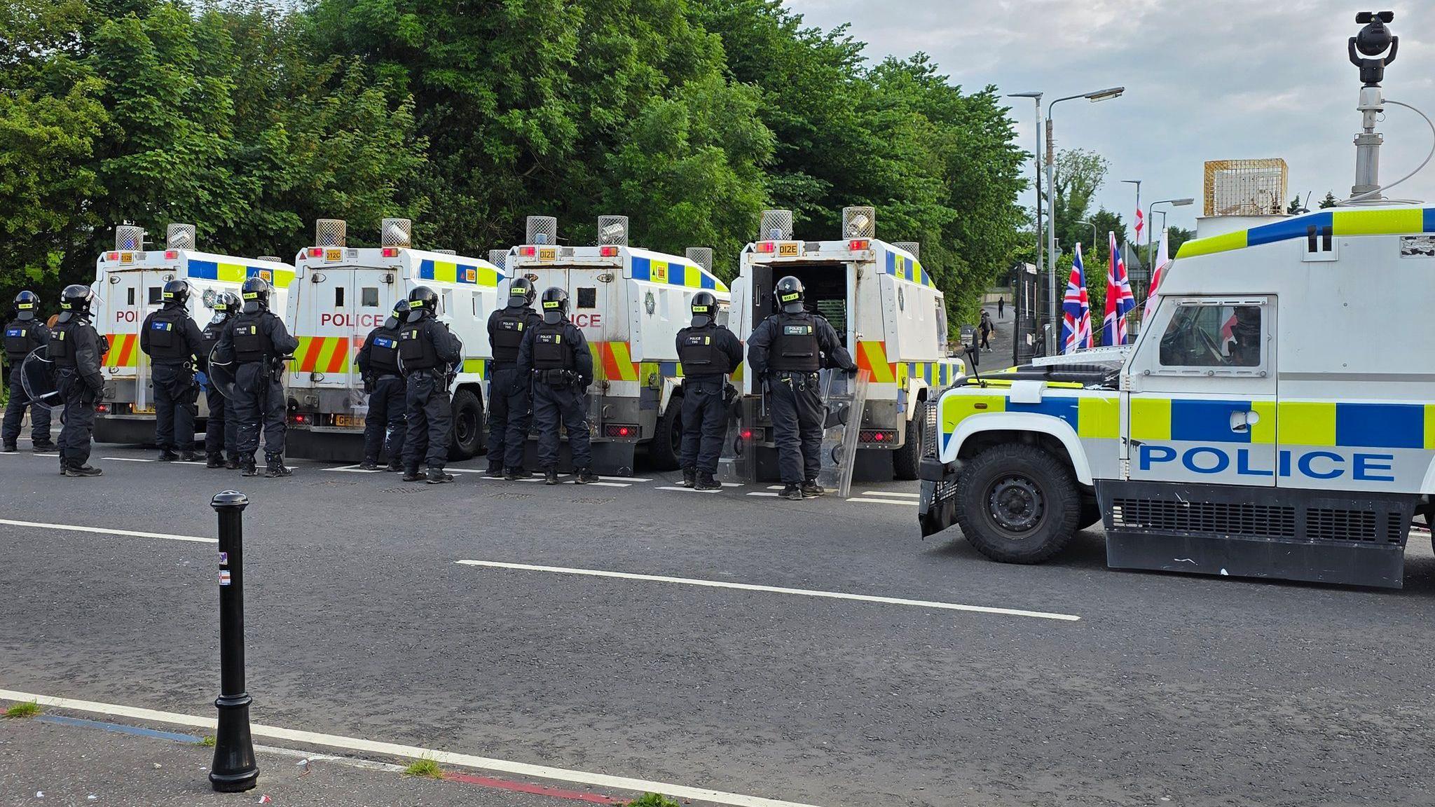 Five police riot vans with a line of police officers behind the. Trees are visible in this distance.