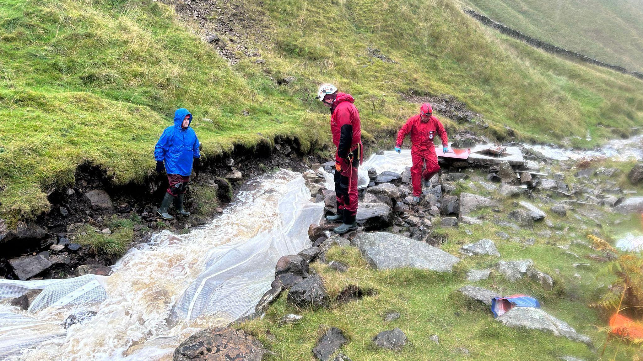 Three people dressed in outdoor gear standing beside a small, fast-flowing stream in a hilly, grassy landscape. The individual on the left is wearing a blue jacket and is positioned at the edge of the stream, possibly observing or preparing to cross. The other two individuals are dressed in red outfits; one is standing on rocks close to the water, while the other is slightly further back, also near the stream.
The surrounding terrain is a mix of green grass and scattered rocks, with hills rising in the background. The sky is overcast, suggesting cloudy or rainy weather conditions.