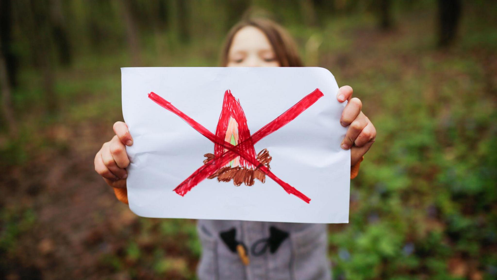 A girl holds up a white piece of paper with a hand drawn camp fire and a big red cross through it