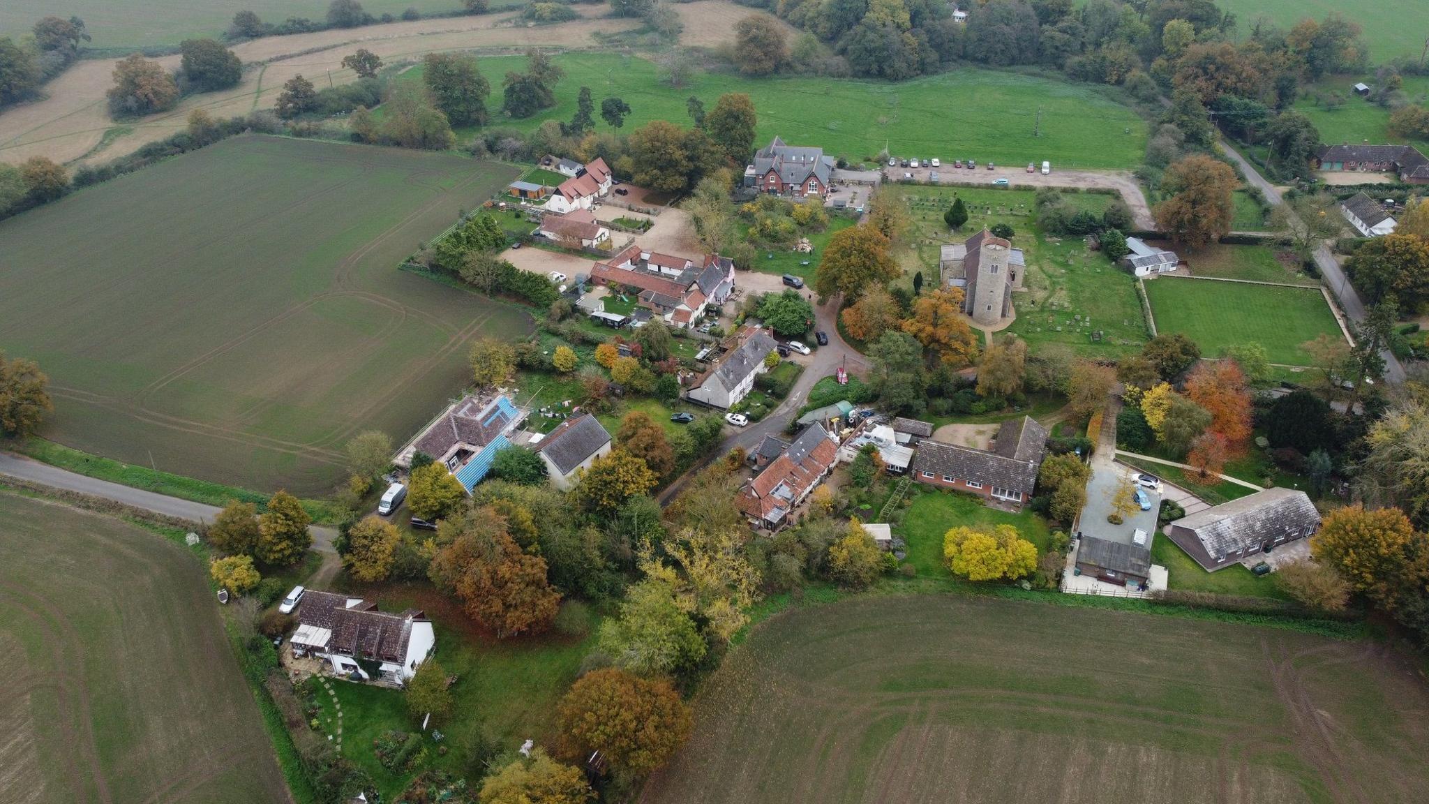 Aerial view of the village of Gissing, surrounded by fields and a large cluster of farm buildings including some houses and farm sheds as well as the parish church.