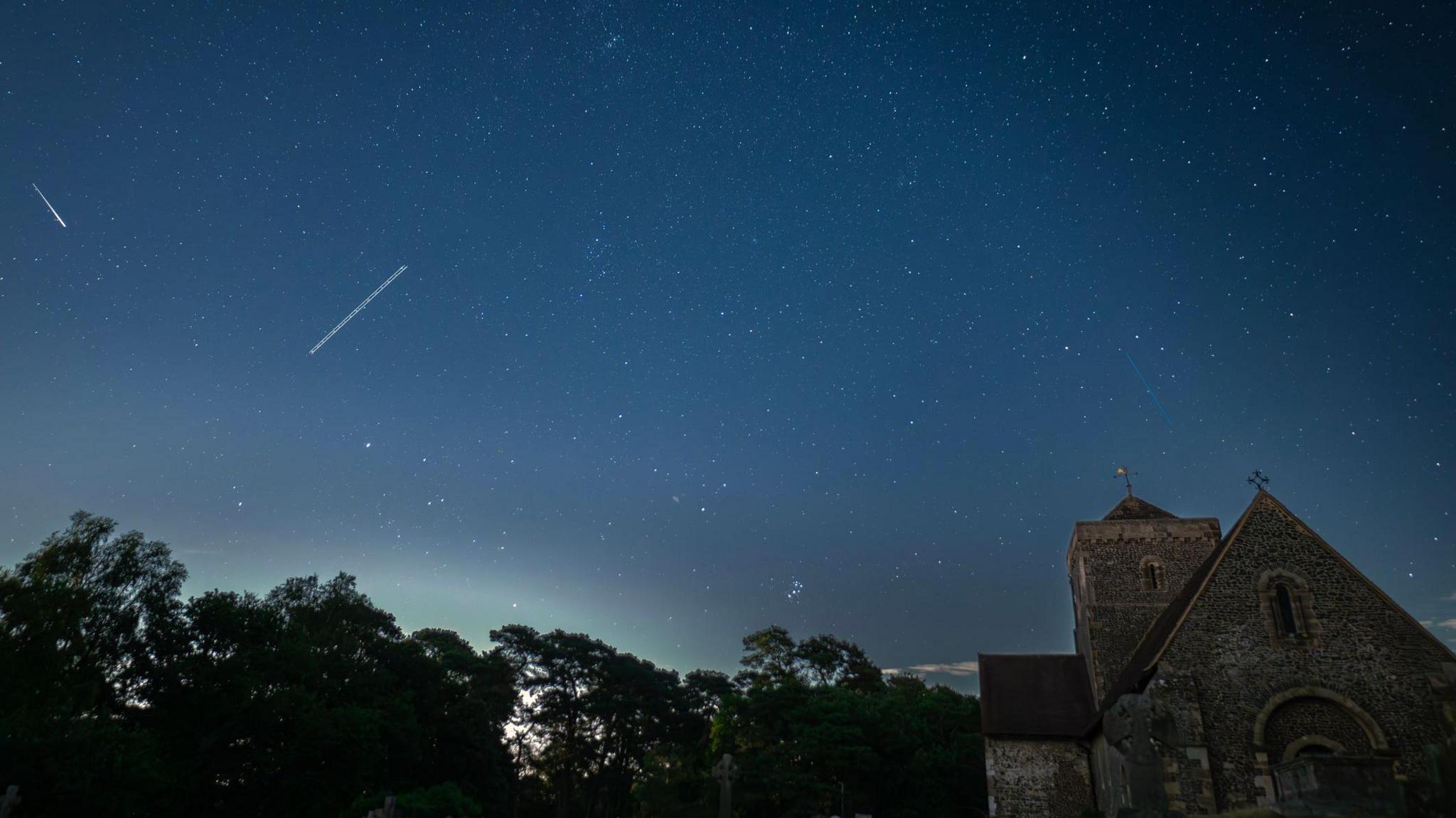 A tranquil nightscape capturing the starlit sky above an old English church graveyard. The silhouettes of weathered gravestones and the gothic architecture of the church create a serene yet mysterious foreground beneath a clear, star-filled sky. A peaceful fusion of history and the cosmos.