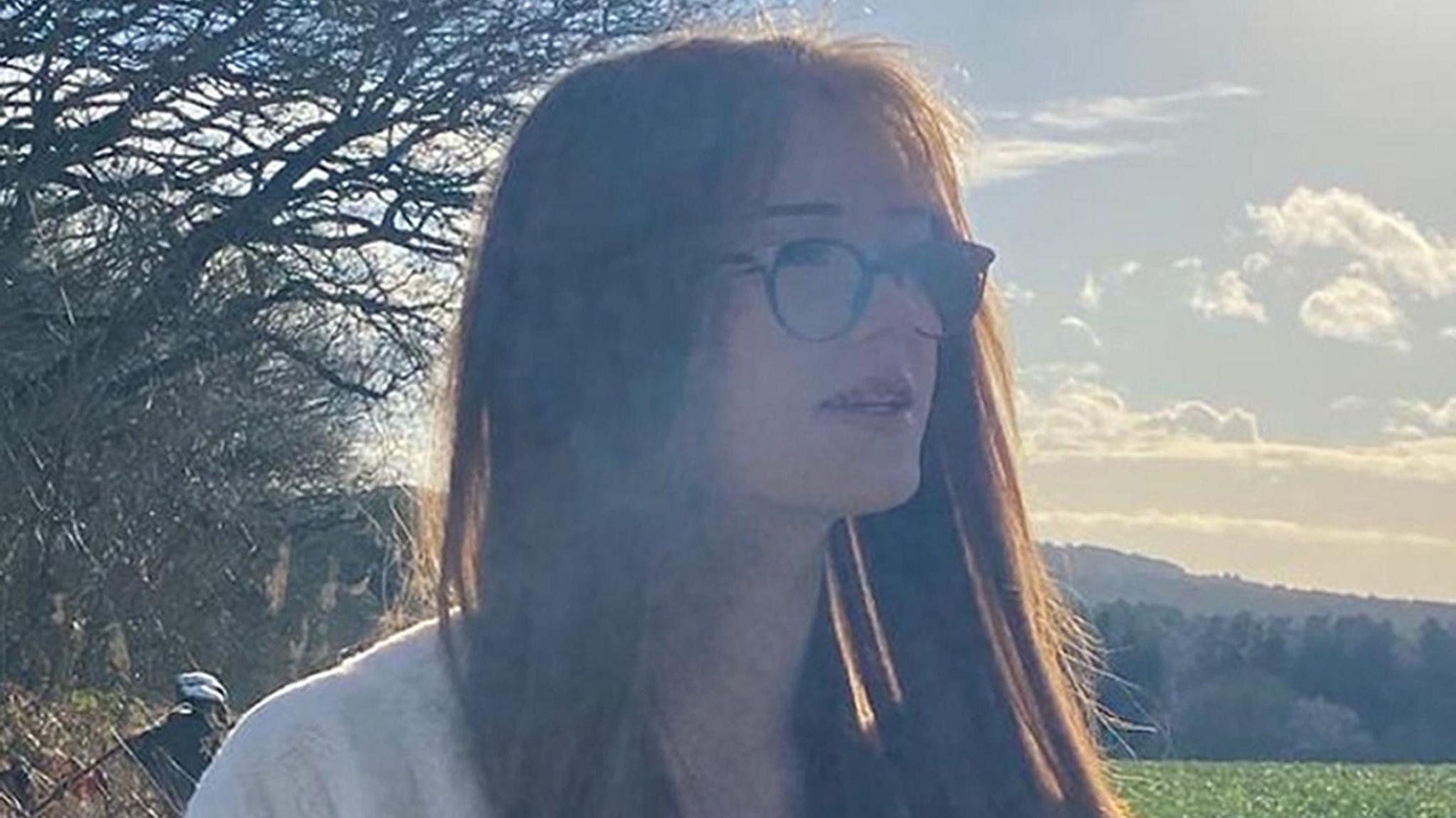 Brianna Ghey staring off in the distance in a park, wearing black framed circular glasses and has long ginger hair