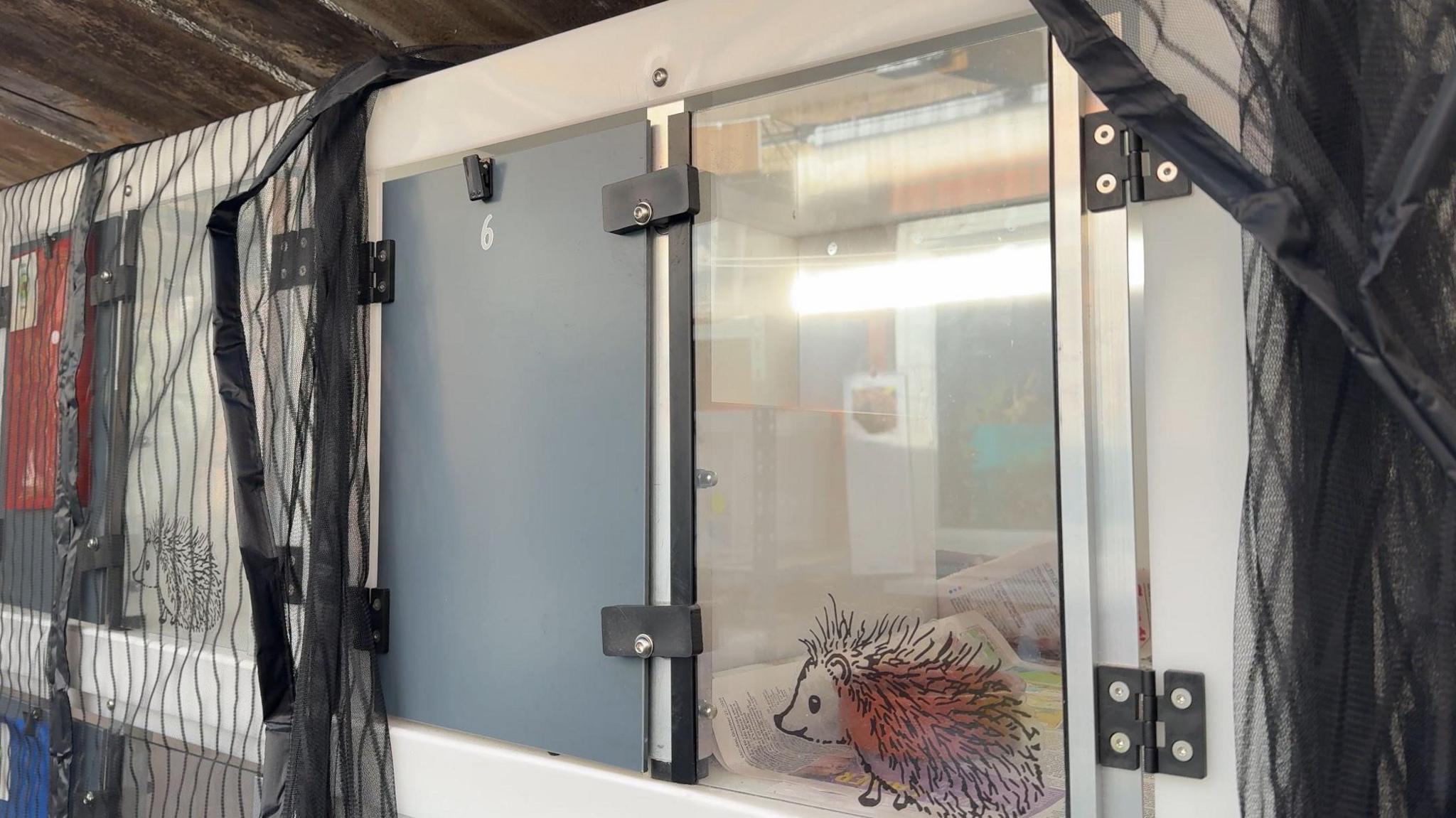 The picture shows what looks like a locker, split into two compartments. One side is a grey door, the other side is clear with a cartoon picture of a hedgehog on. It's part of a stack of similar pods.