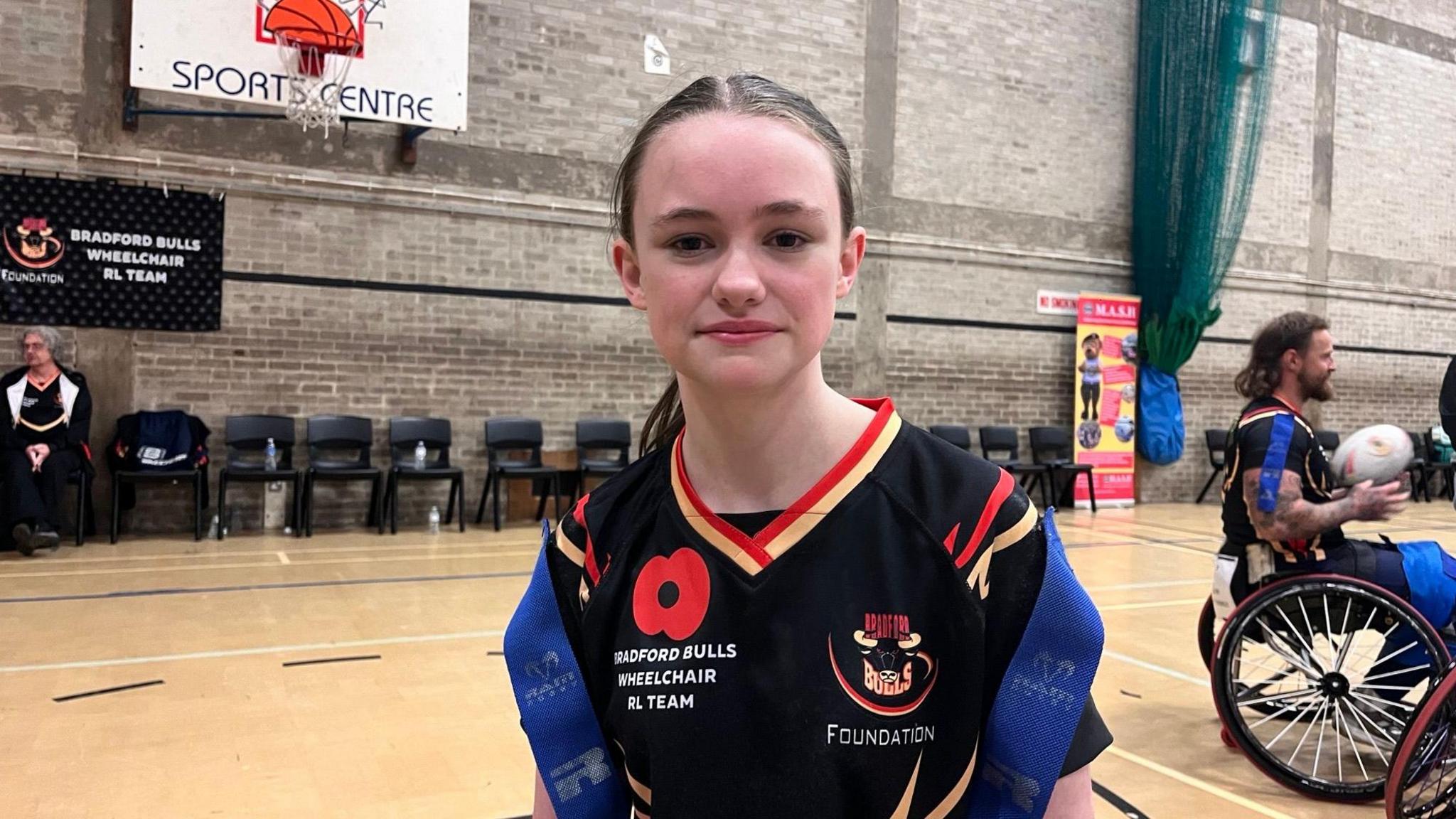 Isabelle looks straight into the camera.  She has her hair tied back and is wearing a black sports top with two blue ribbons on her shoulders.