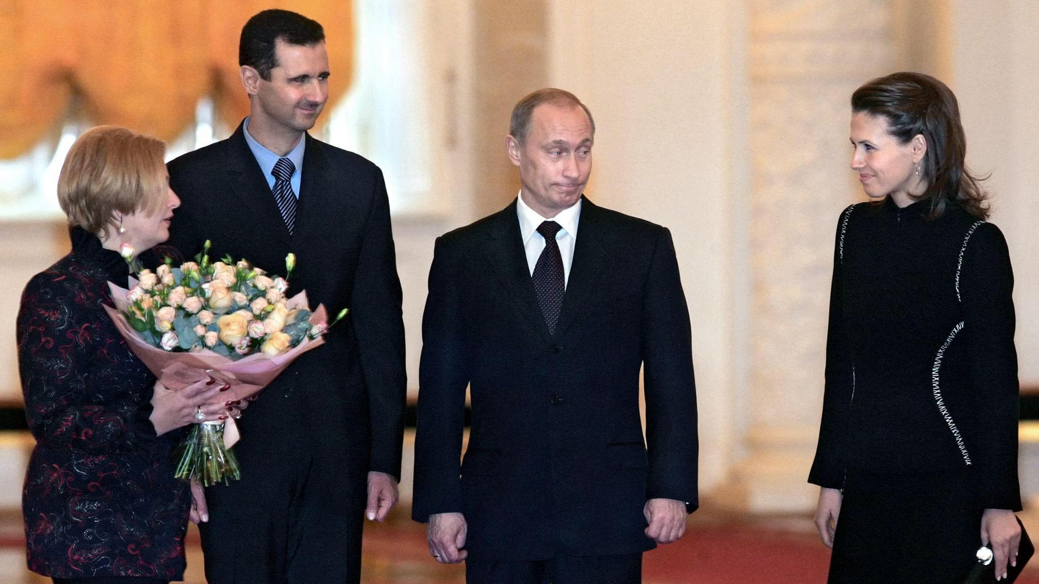 Lyudmila in a patterned black coat holds a bouquet of flowers beside Bashar al-Assad and Vladimir Putin, both in black suits. All three are looking at Asma al-Assad, who wears a dark outfit and carries a purse.