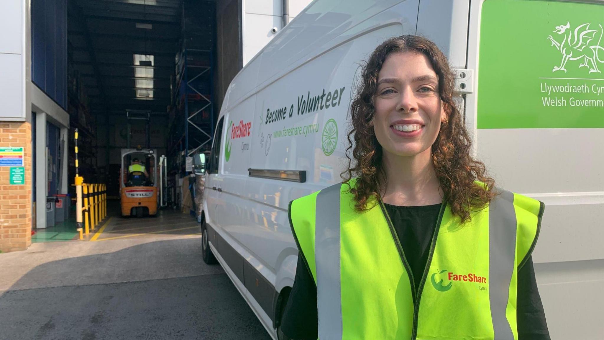 Katie in front of white delivery van by a warehouse in yellow high vis jacket. She has long, dark, curly hair and a forklift truck is visible behind.
